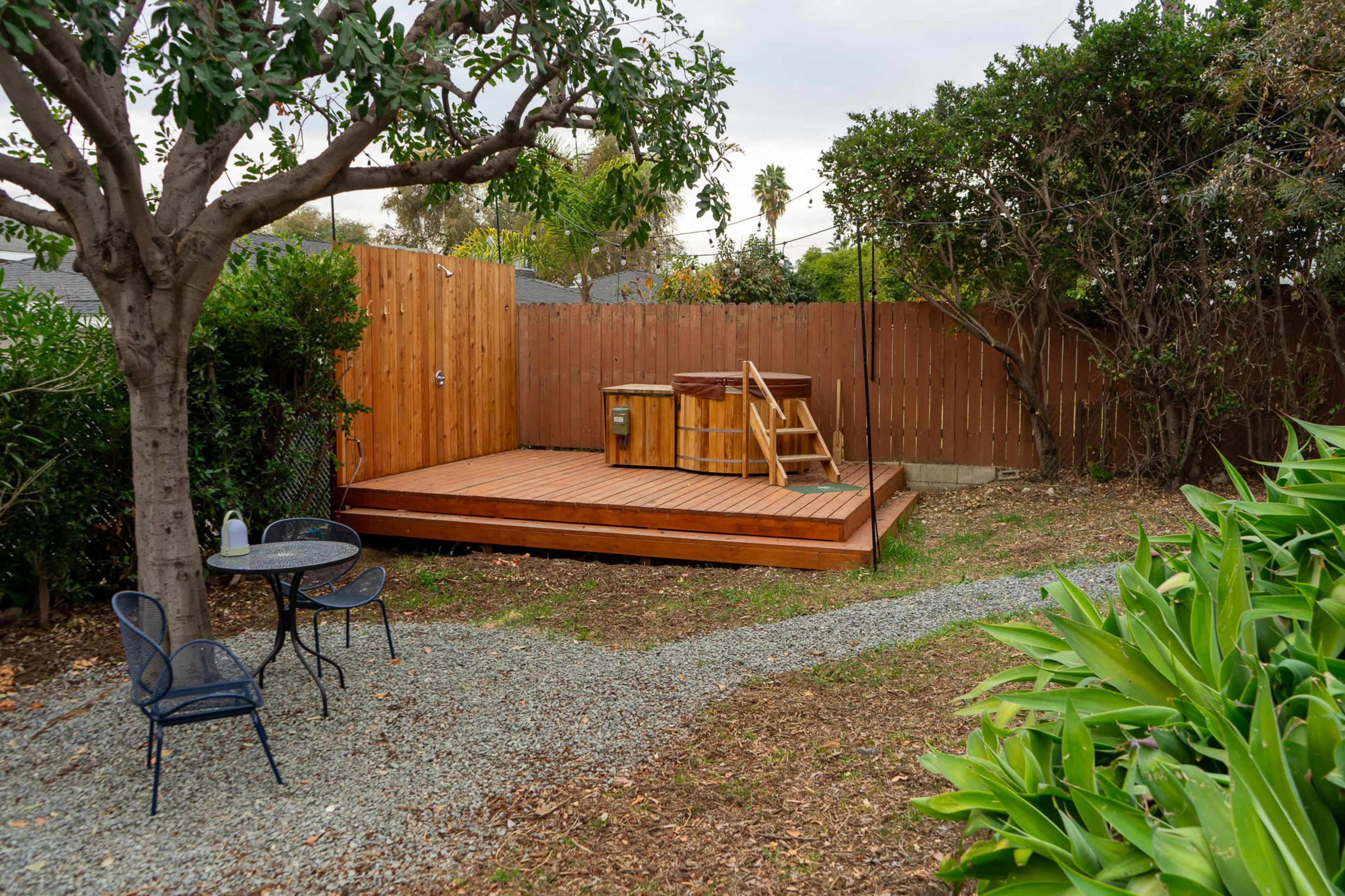 A wooden deck with a hot tub is situated in a landscaped backyard, accompanied by a small table and chairs near a gravel path.