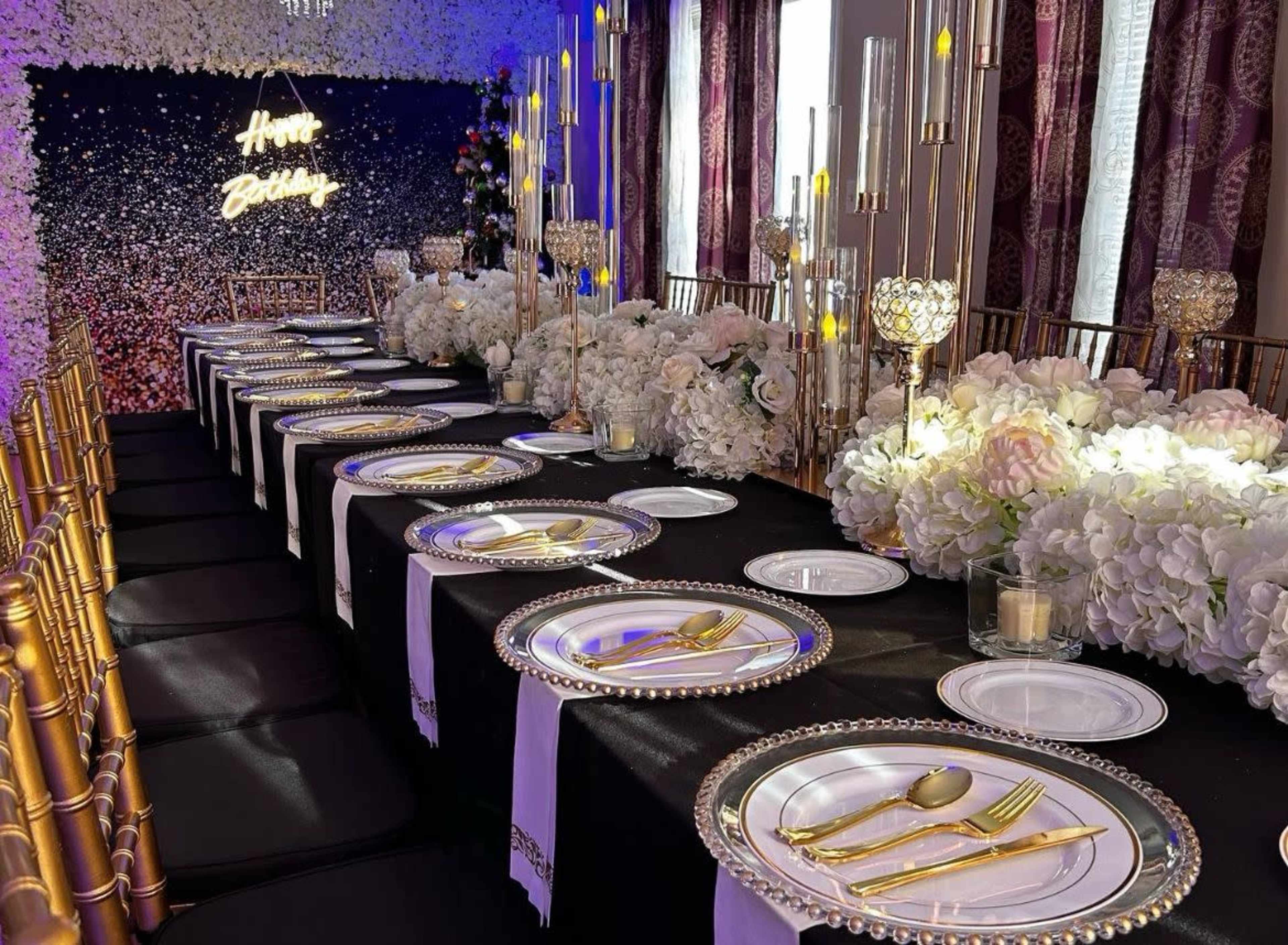 A long dining table is set for a birthday celebration, decorated with white flowers, elegant tableware, and gold accents.