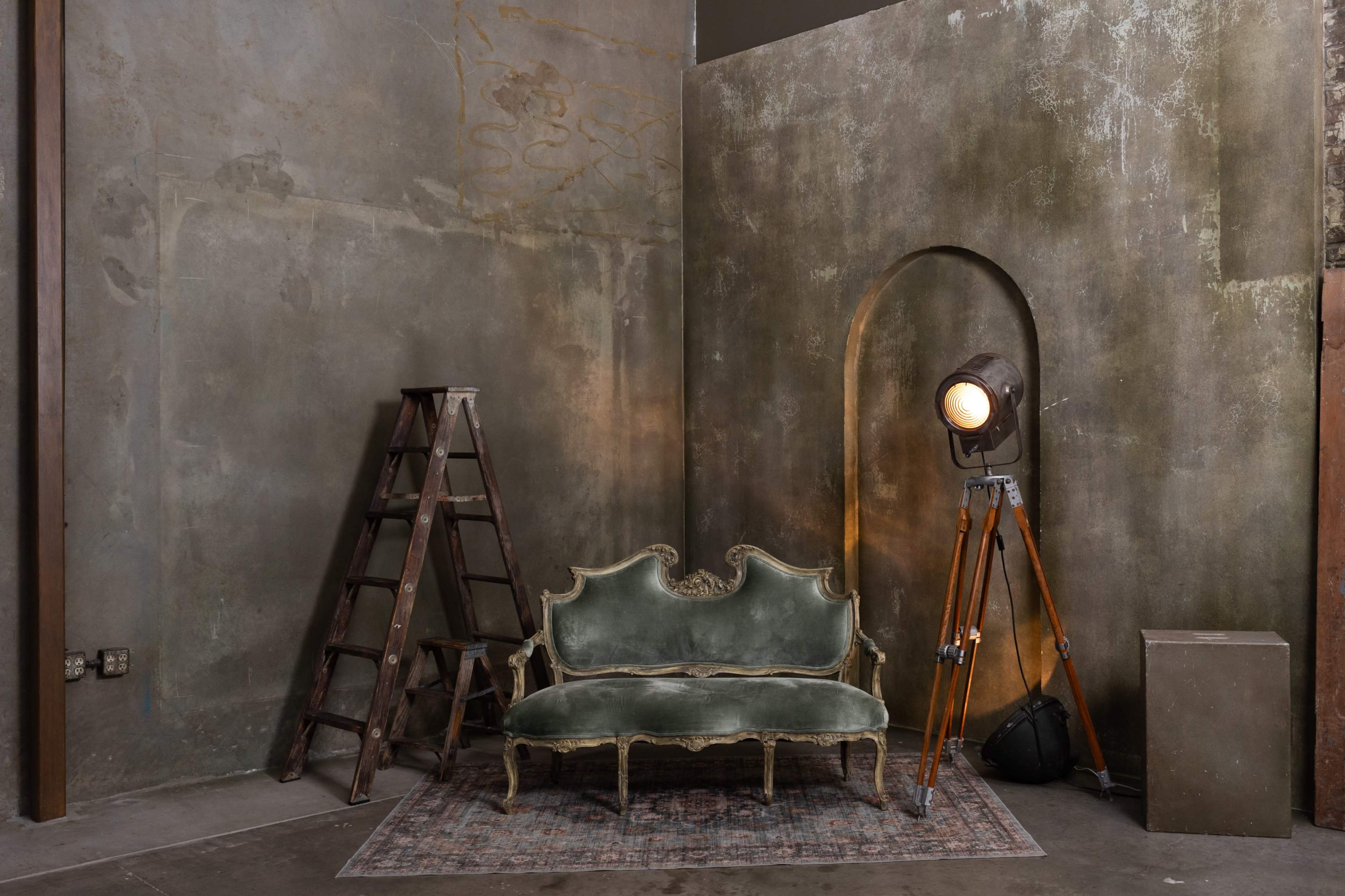 The image shows a vintage green velvet sofa positioned next to a wooden ladder and a large spotlight on a tripod in an unfinished room with textured walls.
