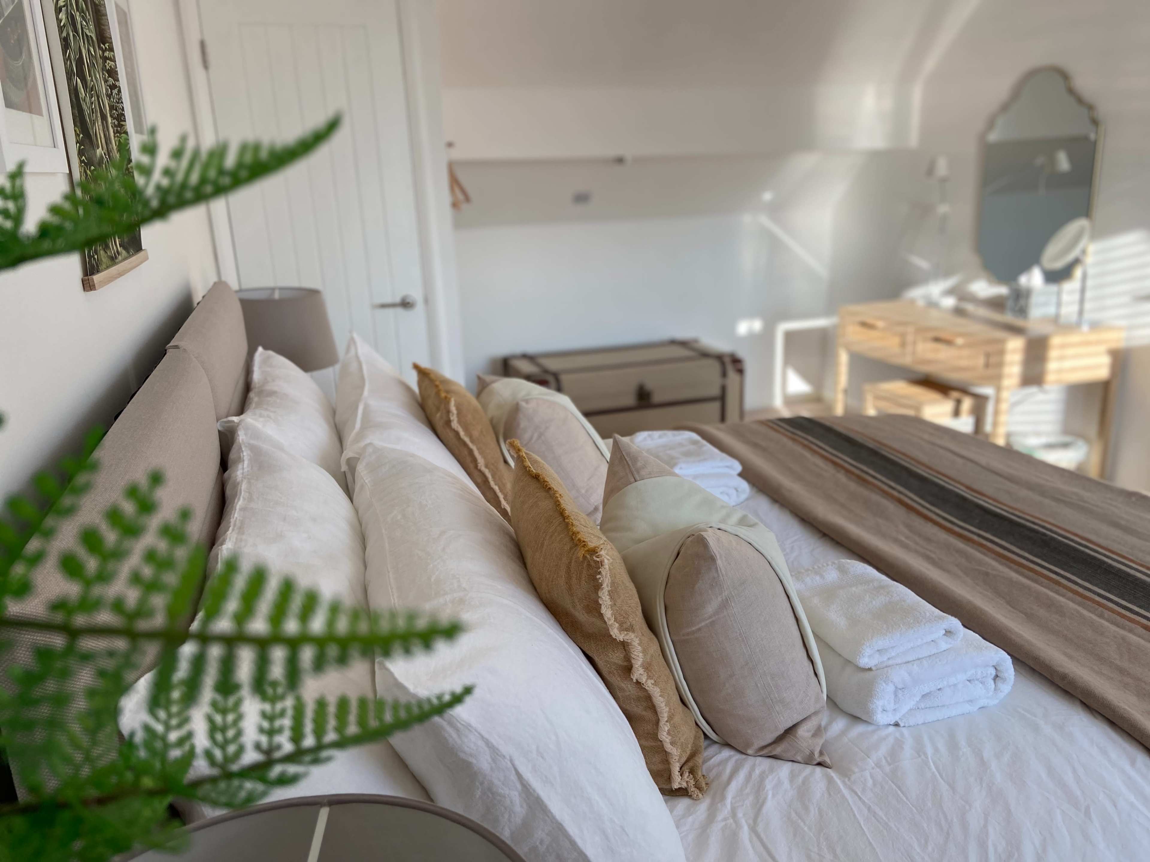 A neatly arranged bedroom features a bed with multiple pillows, a small dresser, and a trunk in the background.