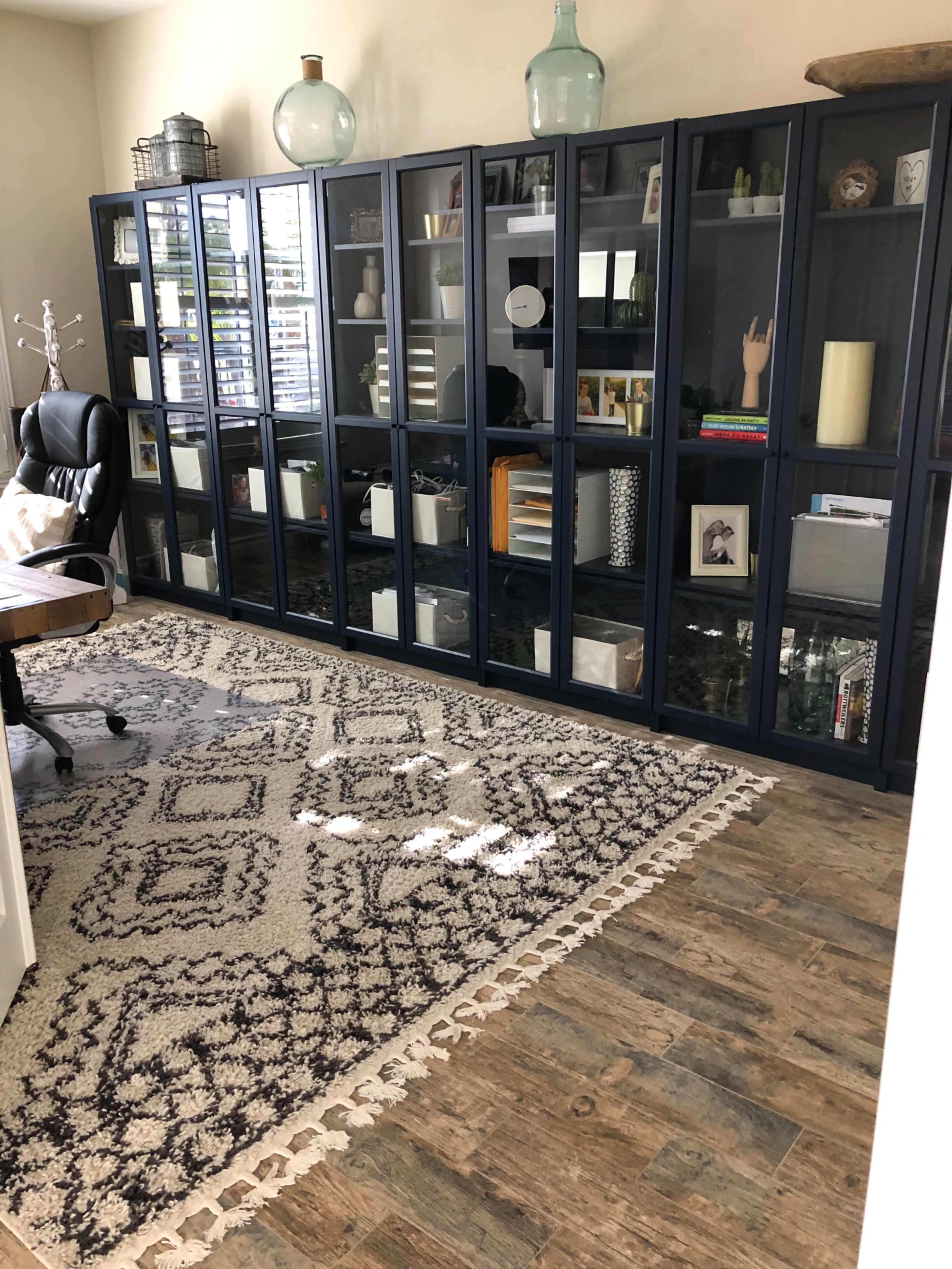 A home office with a wall of dark glass cabinets filled with various decor items, a large patterned rug, and a black office chair at a wooden desk.