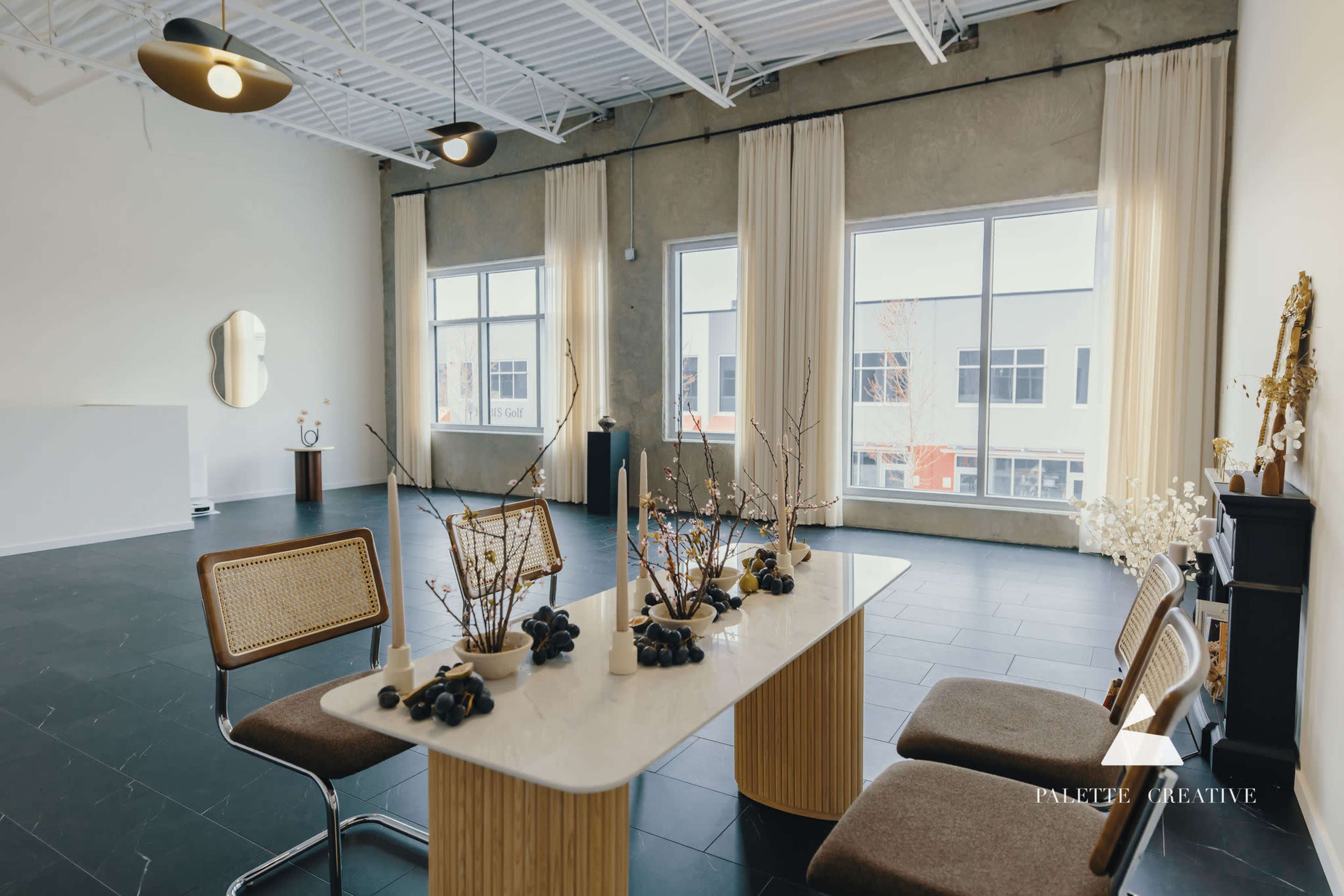 The image shows a modern, spacious room with large windows, a table set with decorative elements, and minimal furnishings.