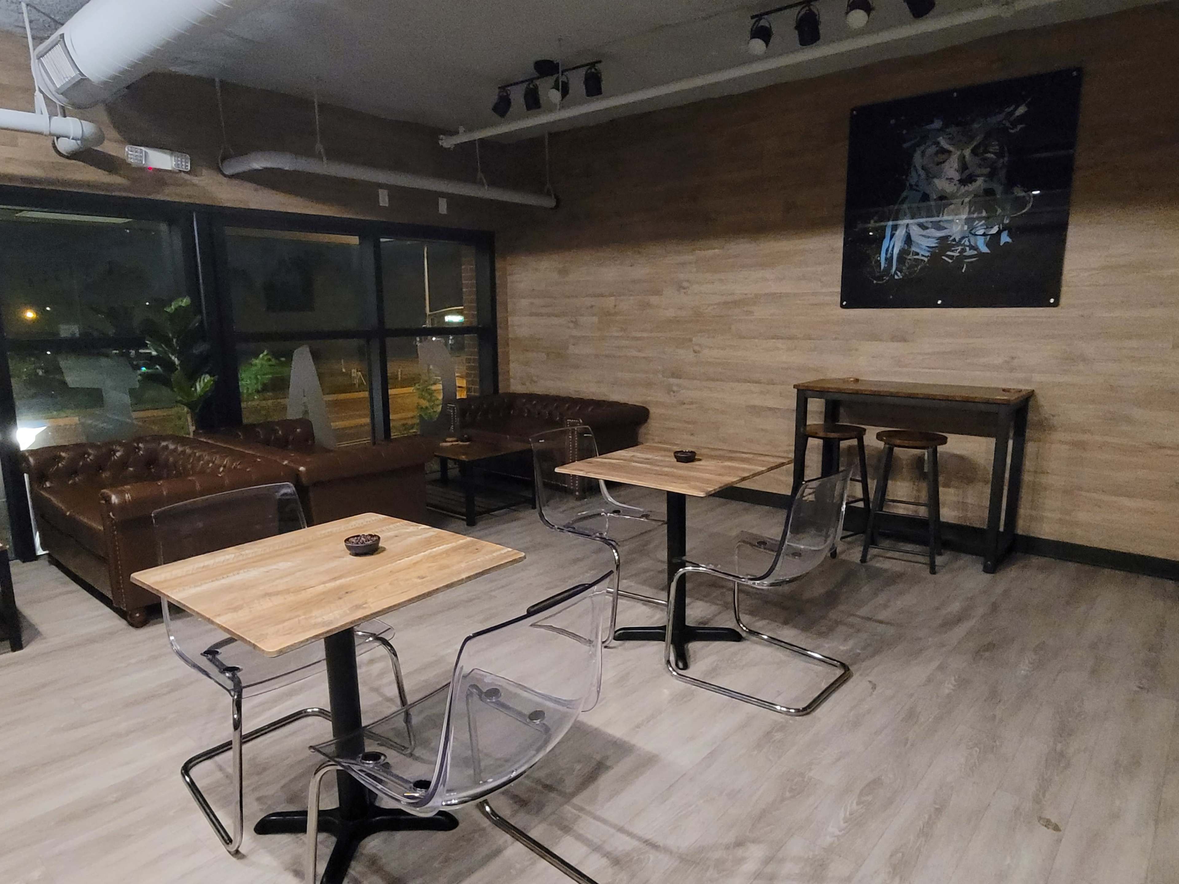 A modern cafe interior with two leather couches, several clear acrylic chairs around wooden tables, and a framed artwork on the wall.