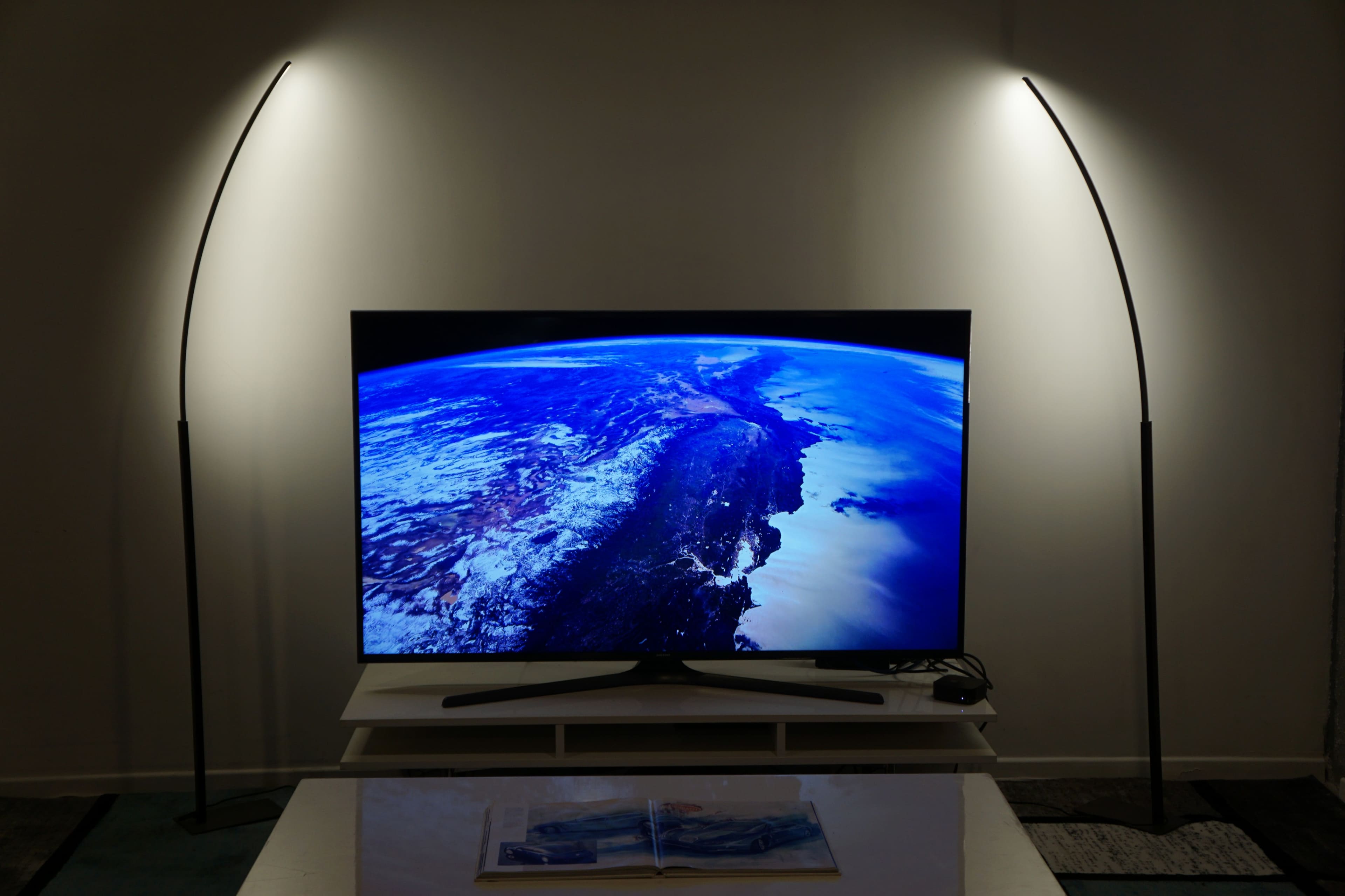 A large television displaying an image of Earth is positioned on a light-colored table, illuminated by two curved floor lamps.