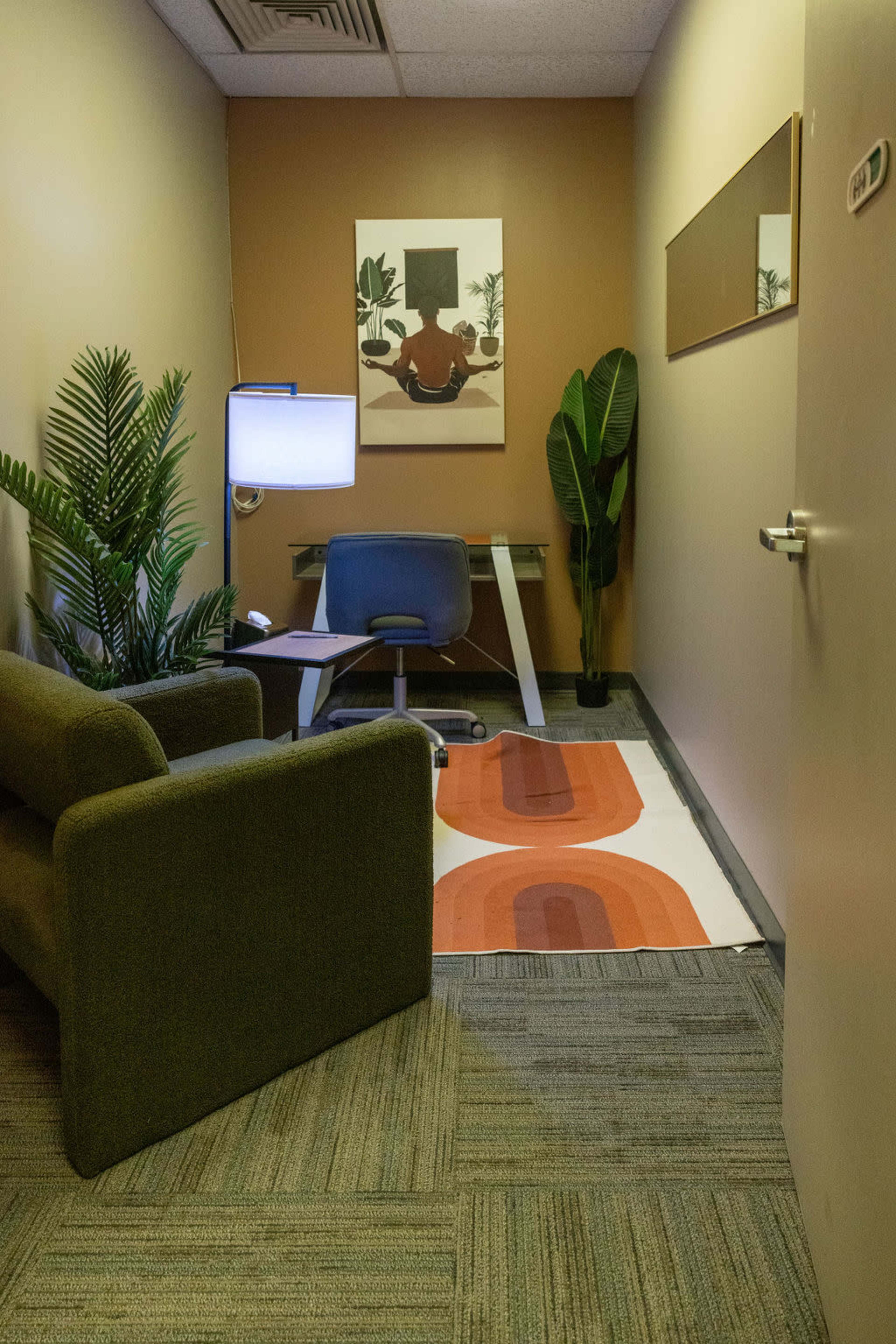 The image shows a small, well-lit office space featuring a sofa, a desk with a chair, a potted plant, and a wall art piece.