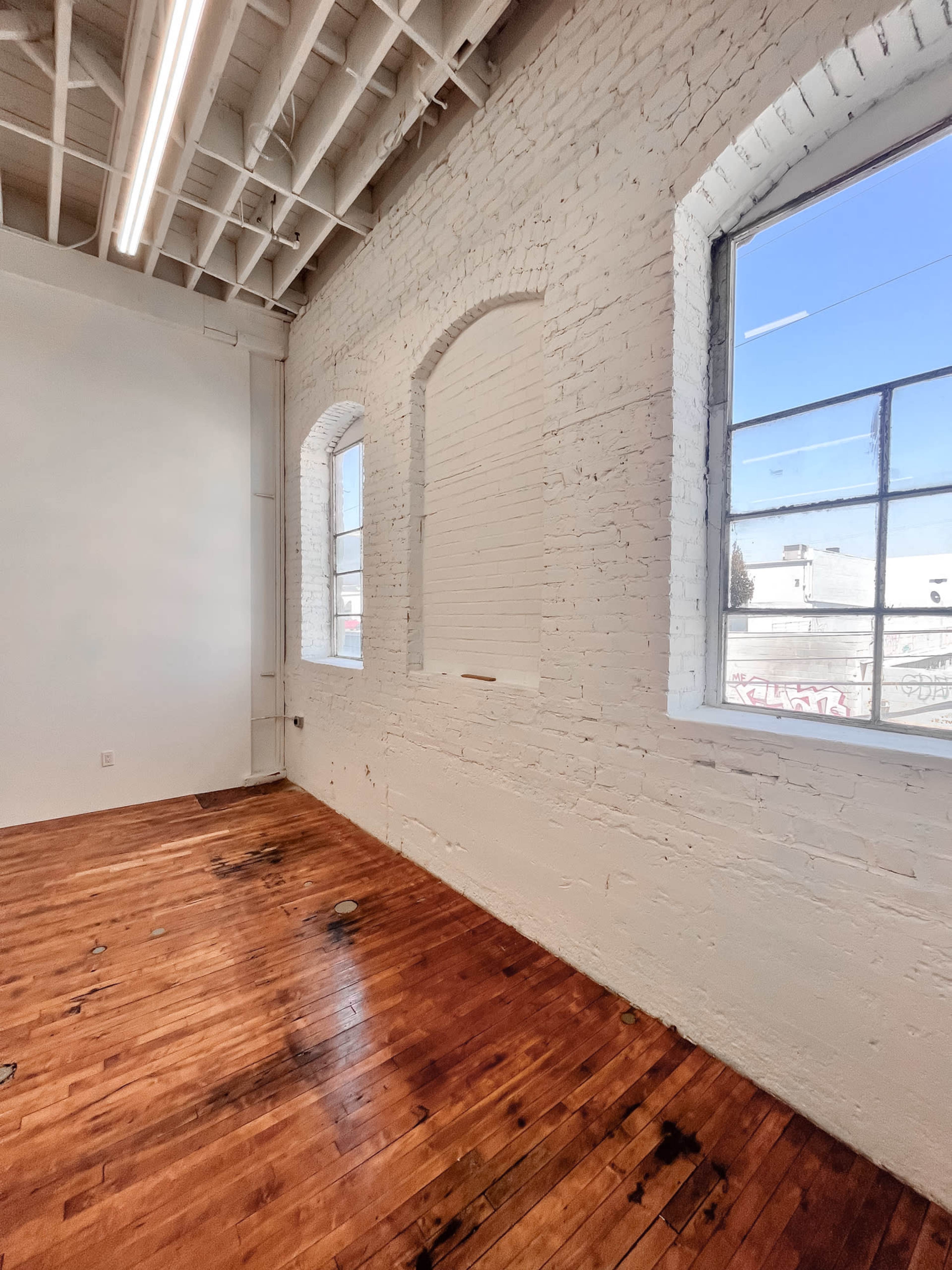 A room with exposed brick walls and two arched windows, featuring hardwood flooring that has visible stains.