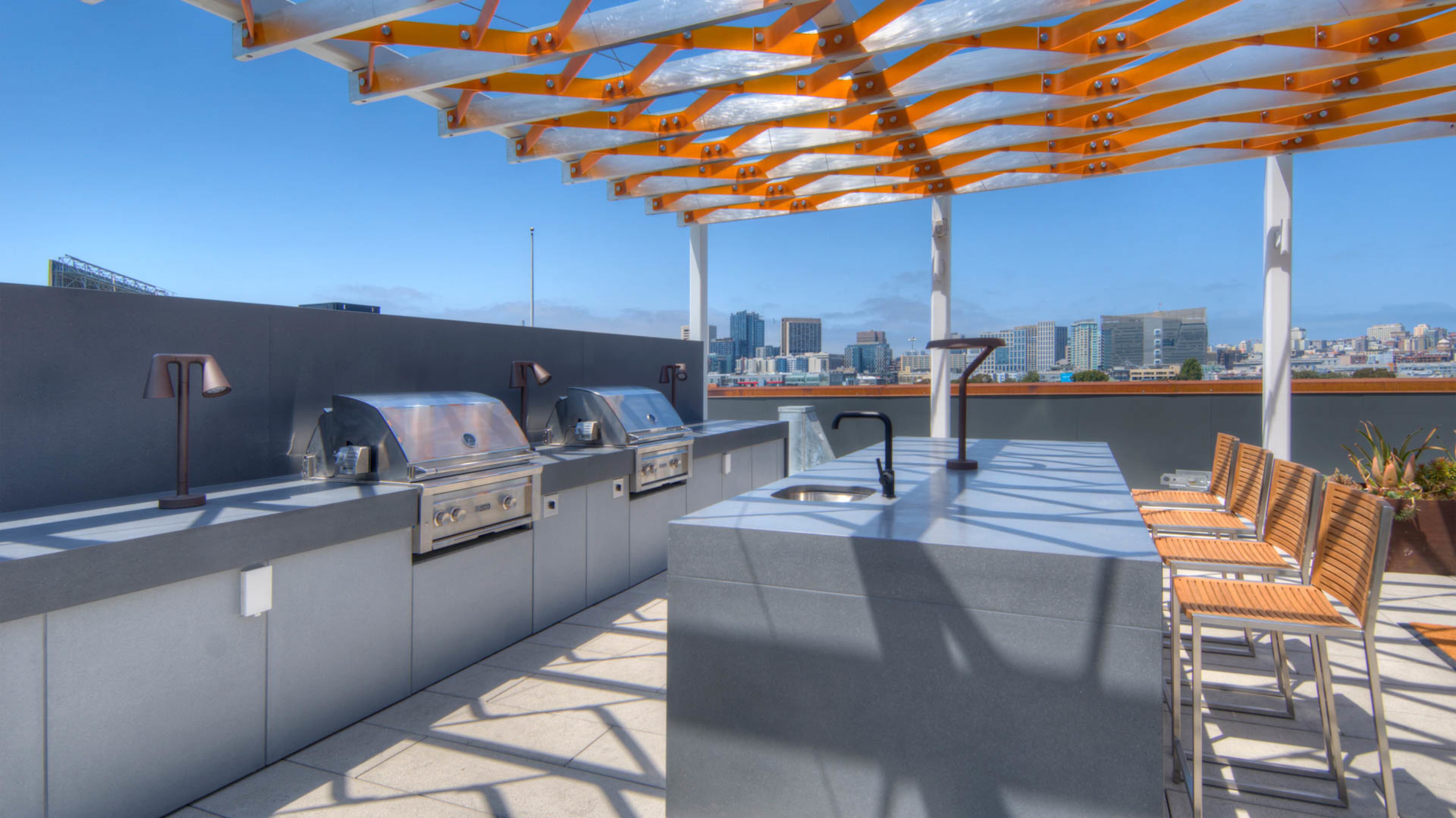 A rooftop outdoor kitchen area featuring stainless steel grills, a sink, and bar seating under a modern pergola with a city skyline in the background.