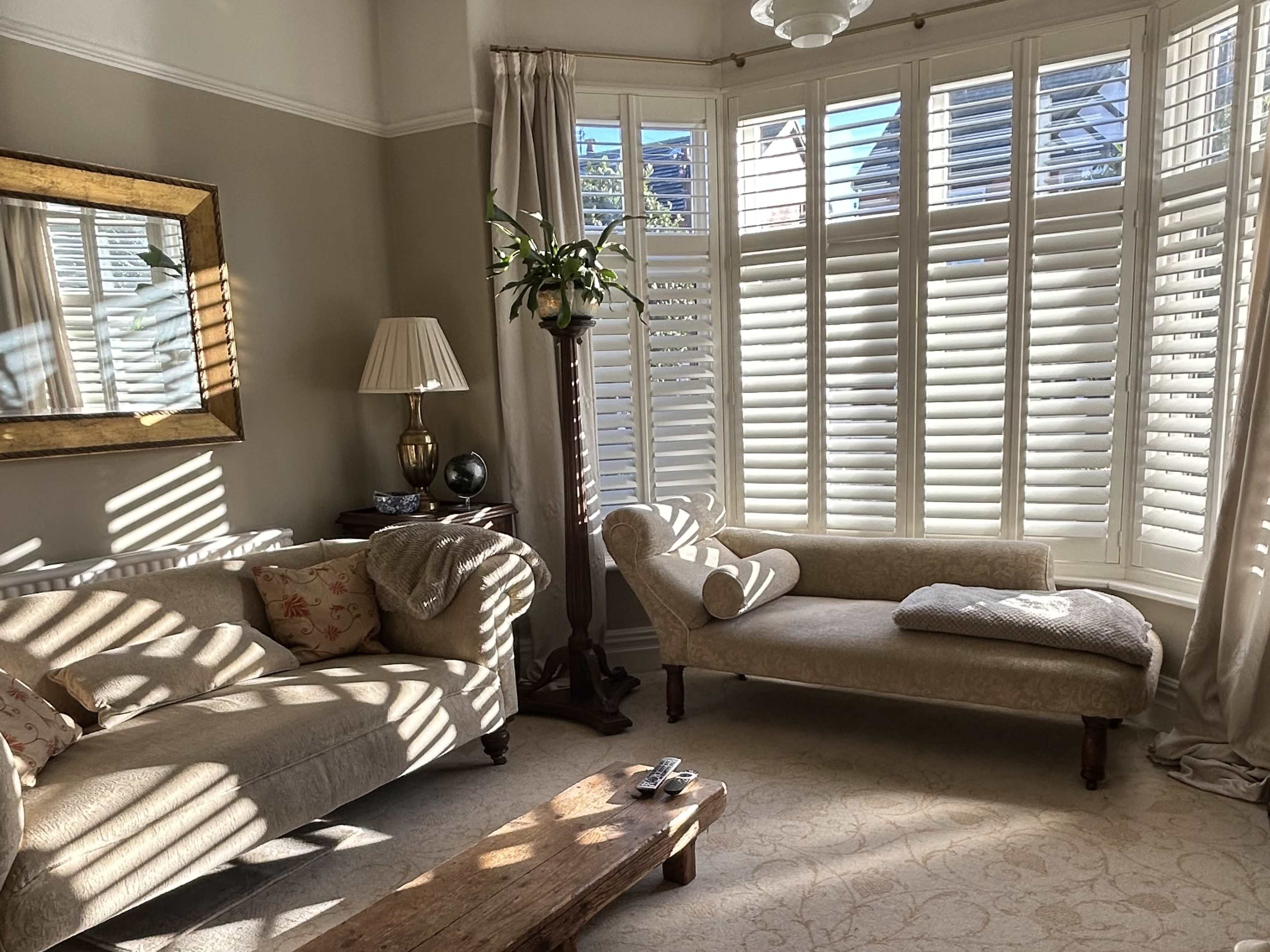 The living room features a beige sofa and a chaise lounge, with sunlight filtering through the window shutters, creating striped patterns on the furniture and floor.