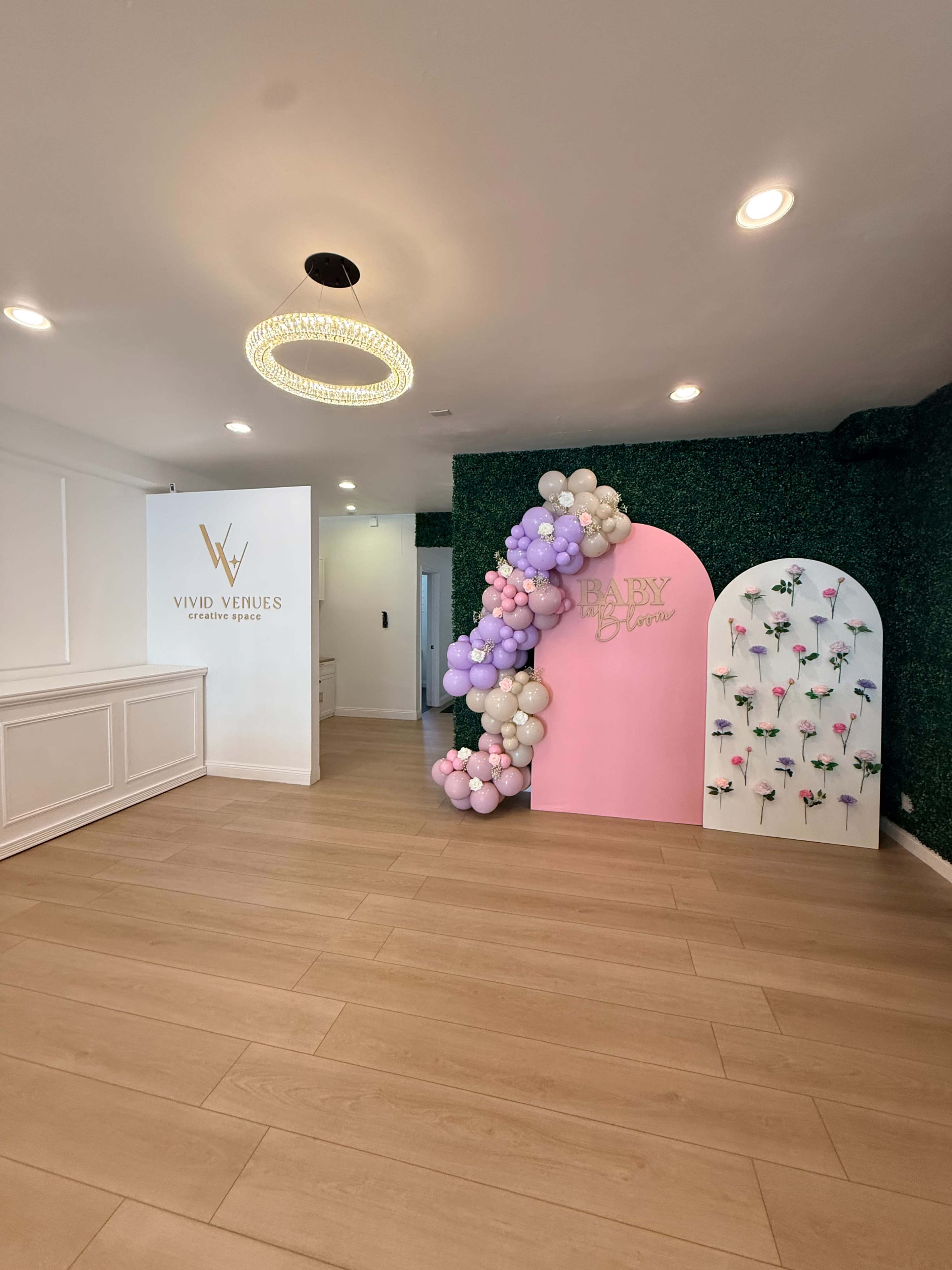 The image shows an indoor event space featuring a decorative wall adorned with balloons in pastel colors and a wall with artificial flowers, alongside a branded wall for "Vivid Venues."