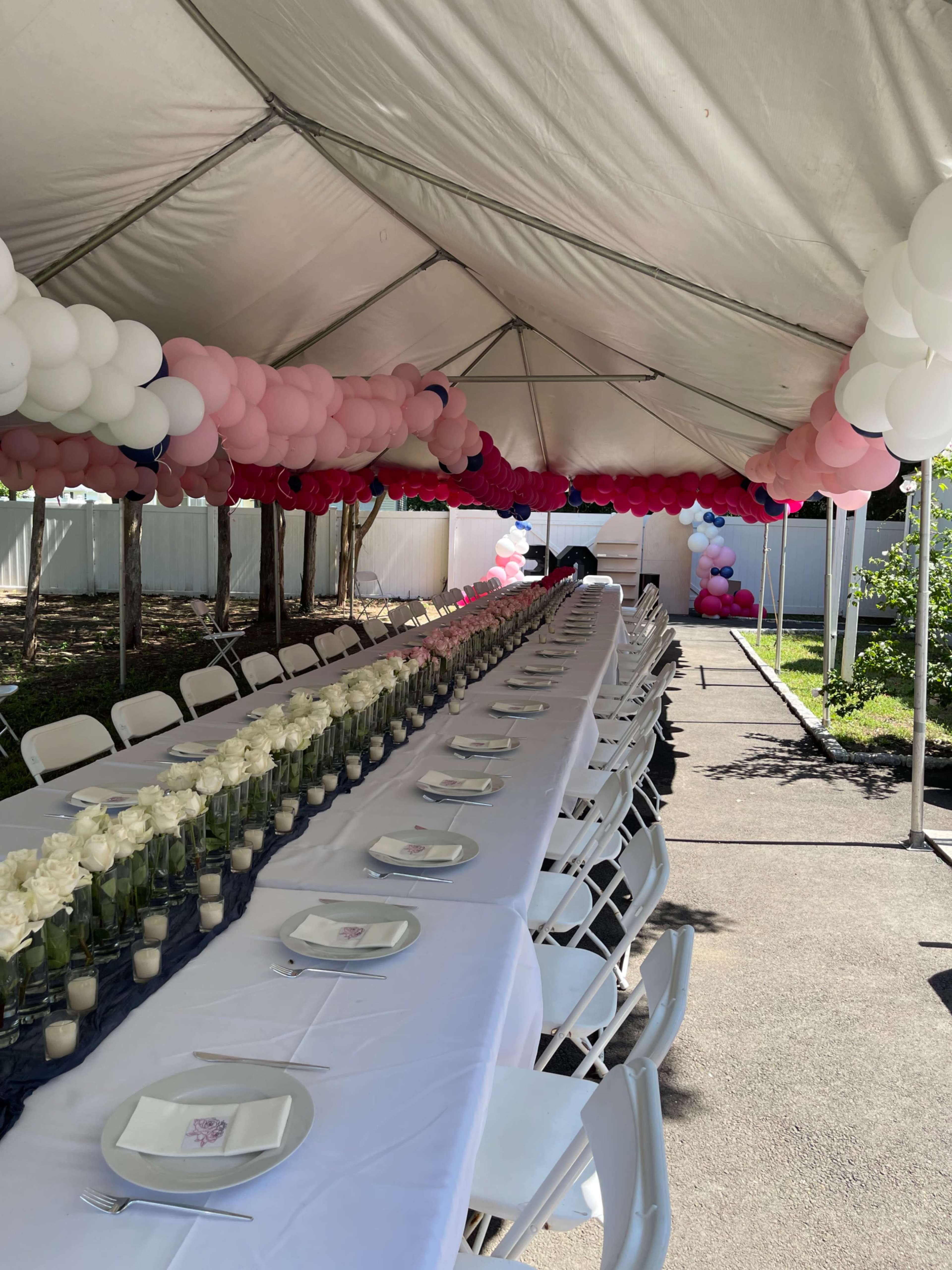A long table is set under a tent, adorned with white roses in glass vases, and pink and white balloons hang above.