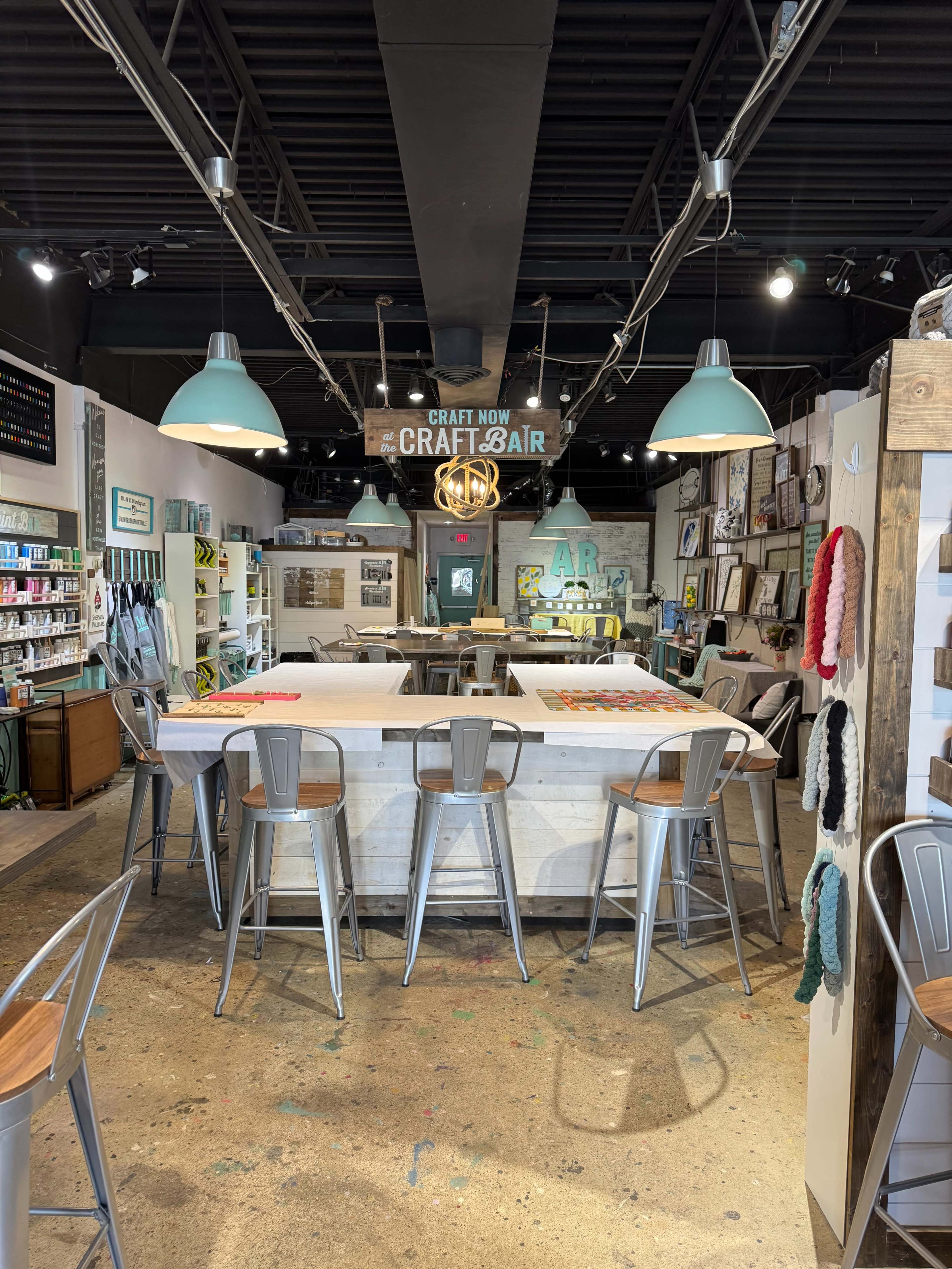 The image shows a craft studio with a white central table surrounded by metal bar stools, shelves filled with craft supplies, and overhead lights.