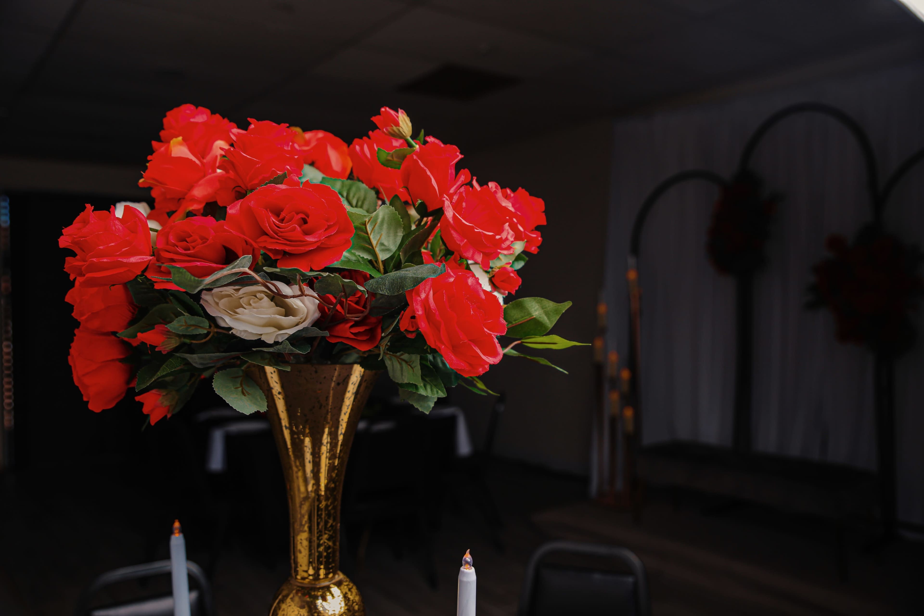 A large bouquet of red and white artificial roses sits in a gold vase on a table in a decorated venue.
