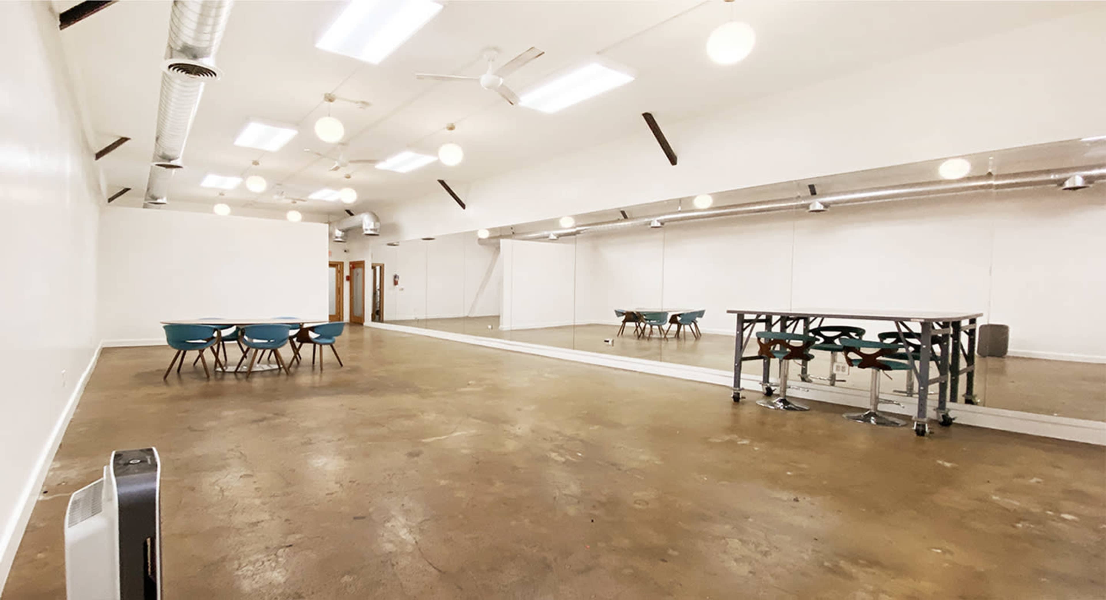 The image shows a spacious, empty room with a polished concrete floor, a mirrored wall, and several tables and chairs arranged in the center.