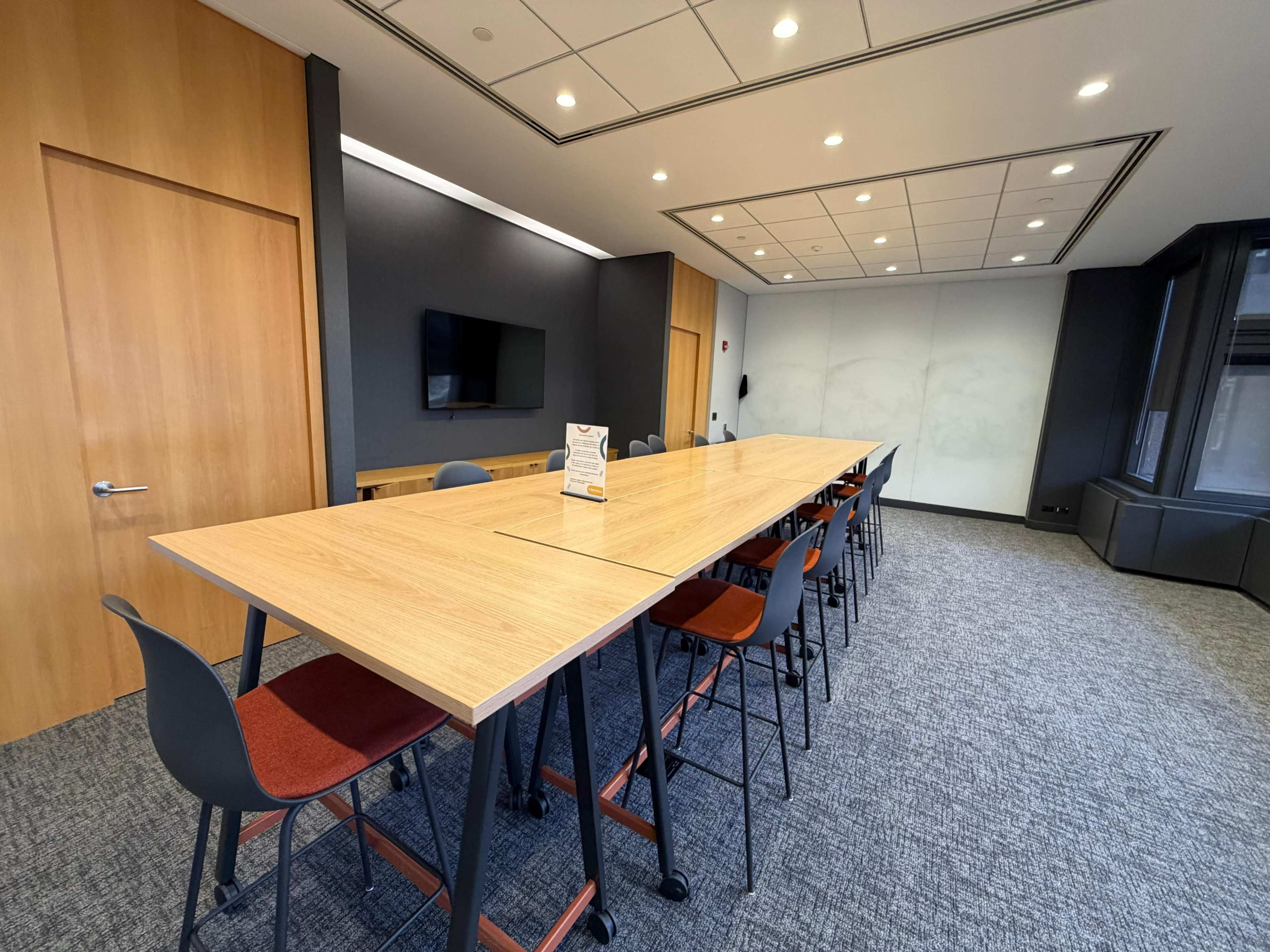 A long conference table with chairs is arranged in a modern meeting room featuring wooden doors, a television, and large windows.