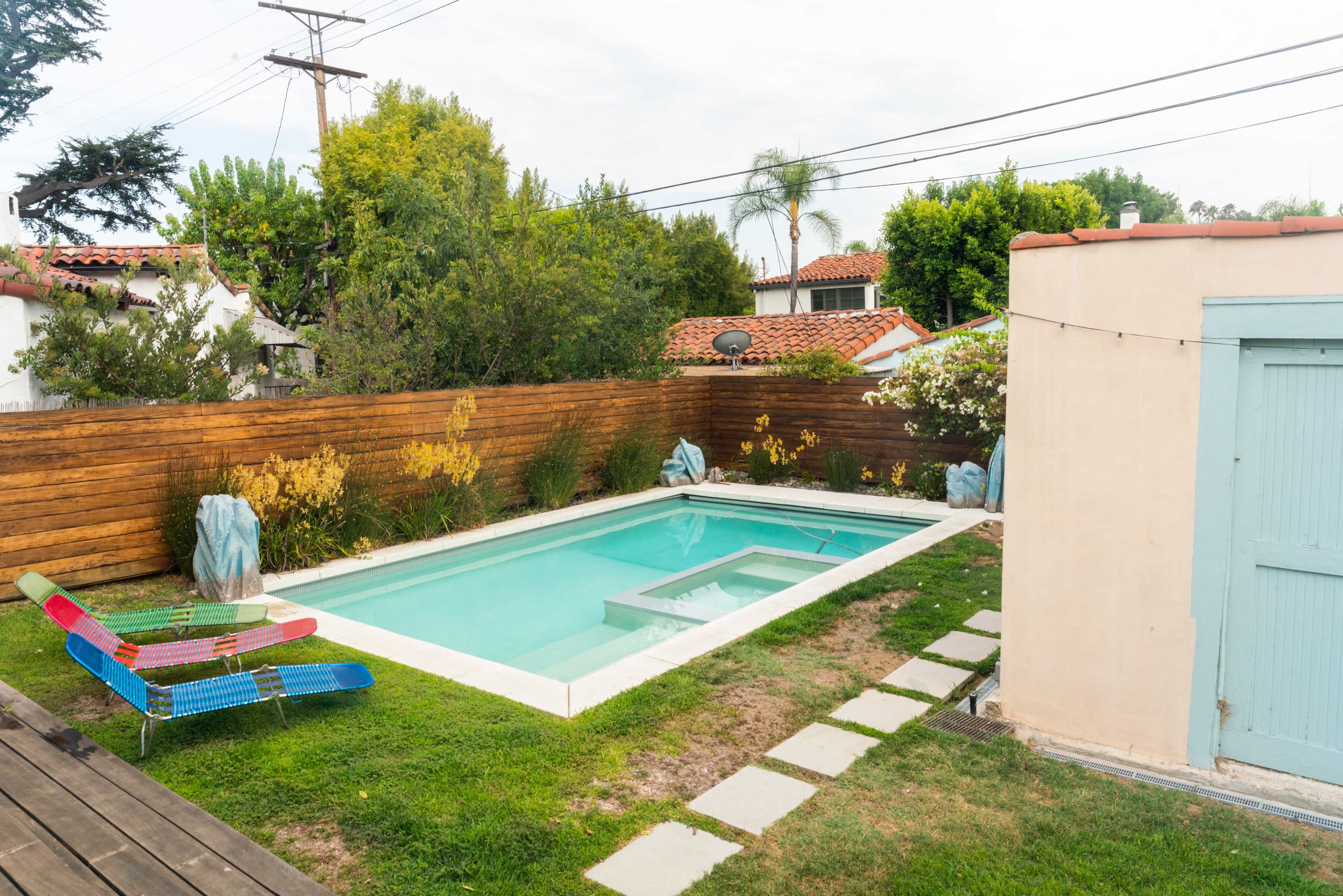 A shallow rectangular pool surrounded by grass and wooden fencing, with a colorful lounge chair and nearby greenery.