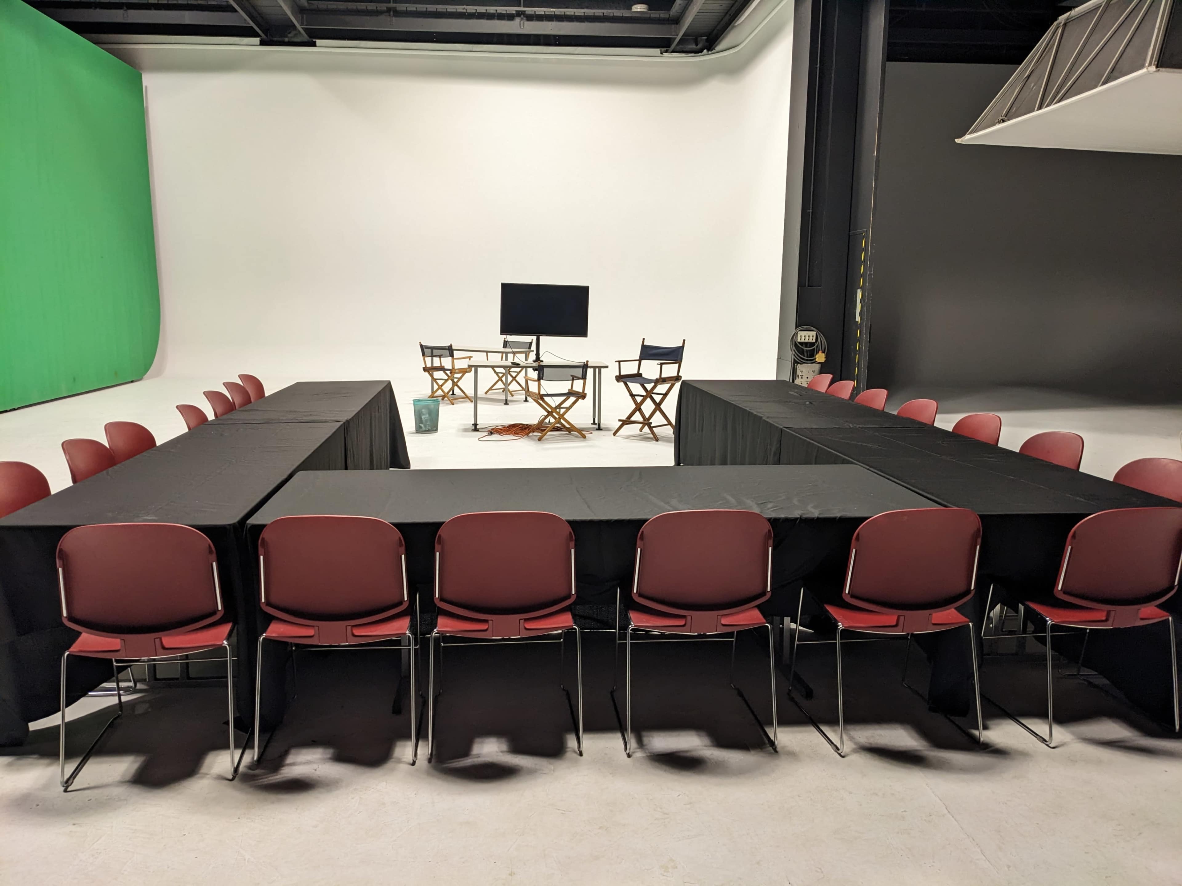 A studio setup features a rectangular arrangement of tables with chairs around them, a screen, and director's chairs at one end.