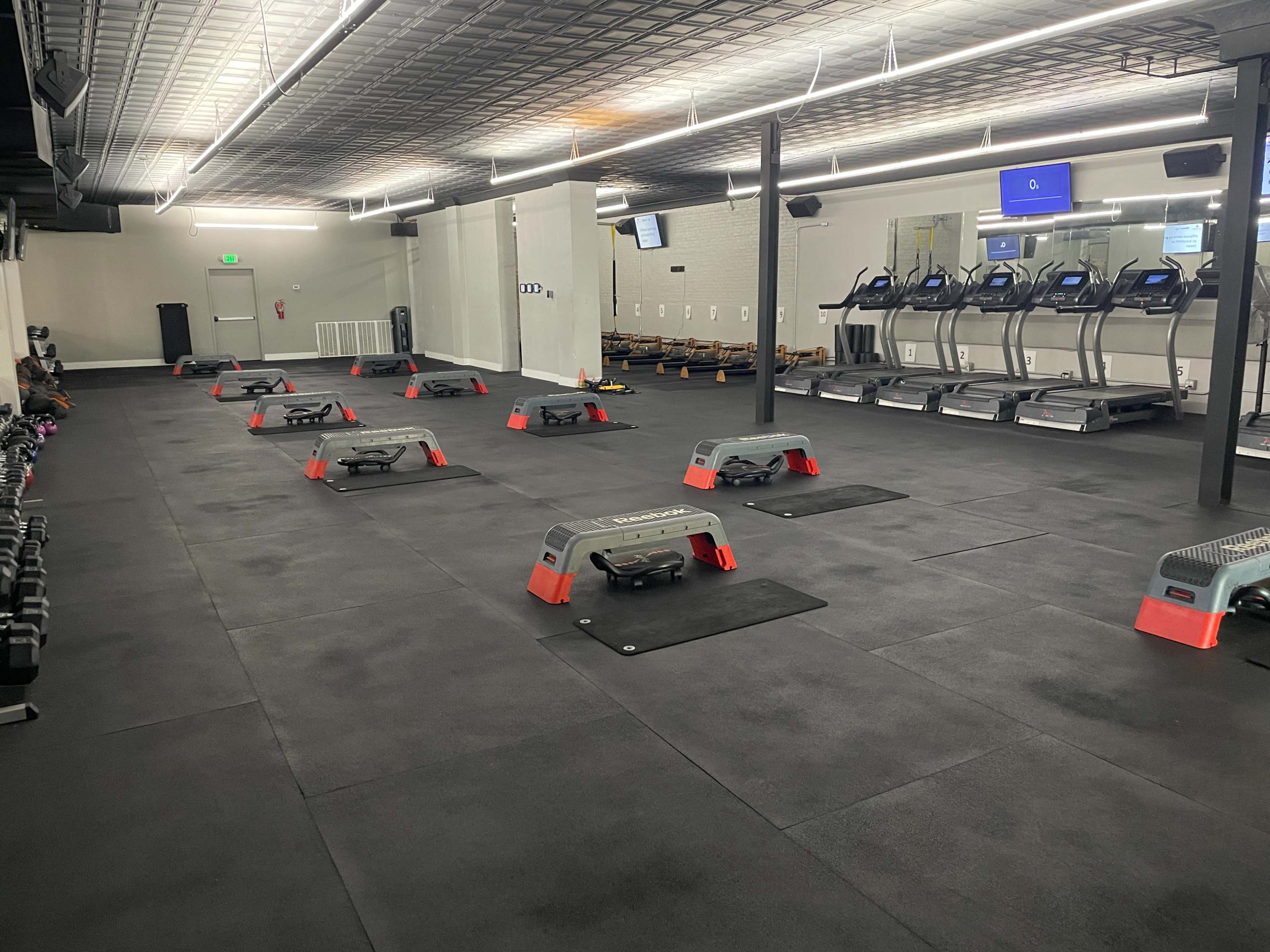 A gym interior with multiple exercise stations featuring step platforms and weights, along with a row of treadmills in the background.