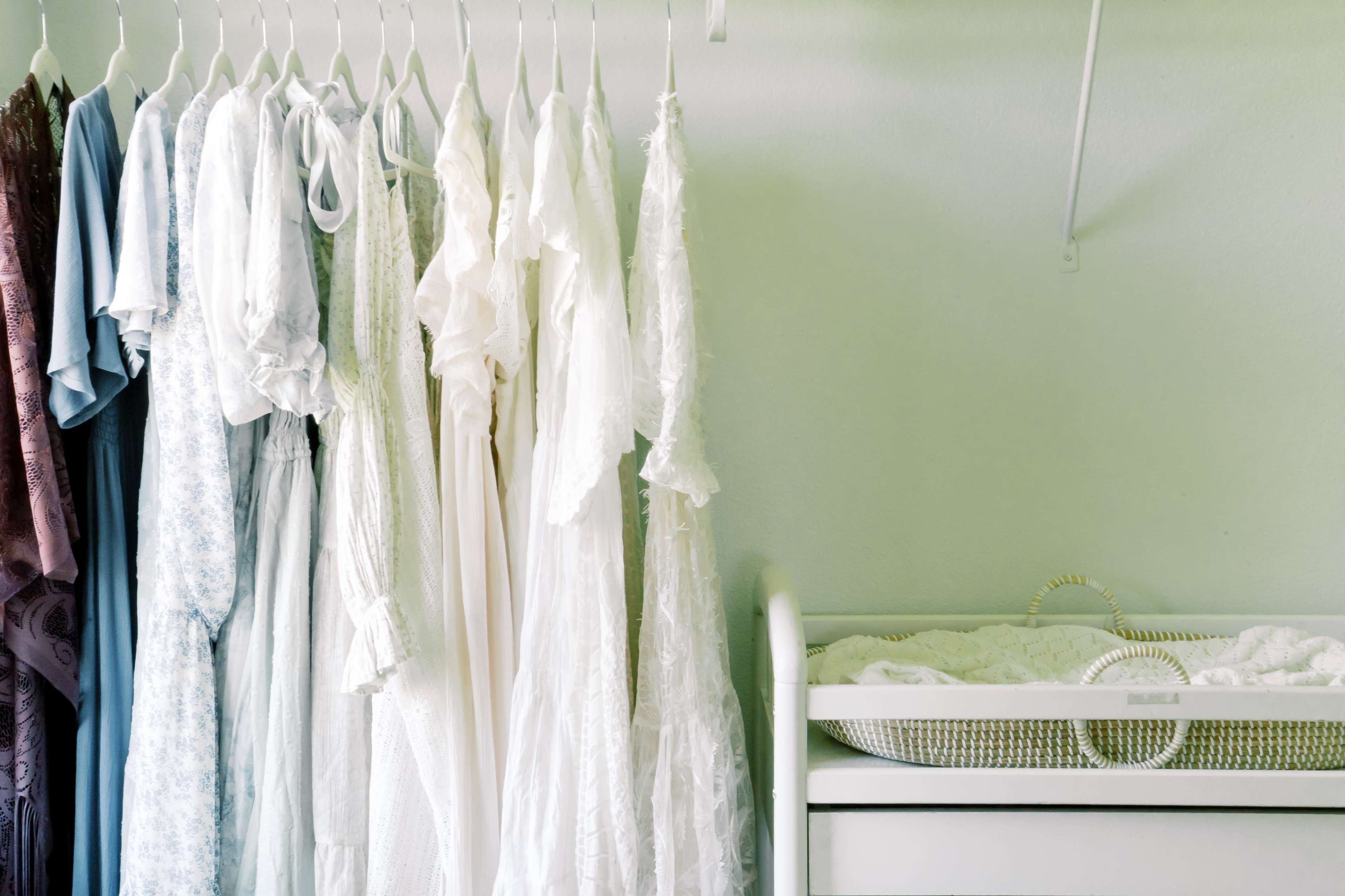A row of hanging clothing in various styles and colors is displayed next to a baby bassinet in a softly lit room.