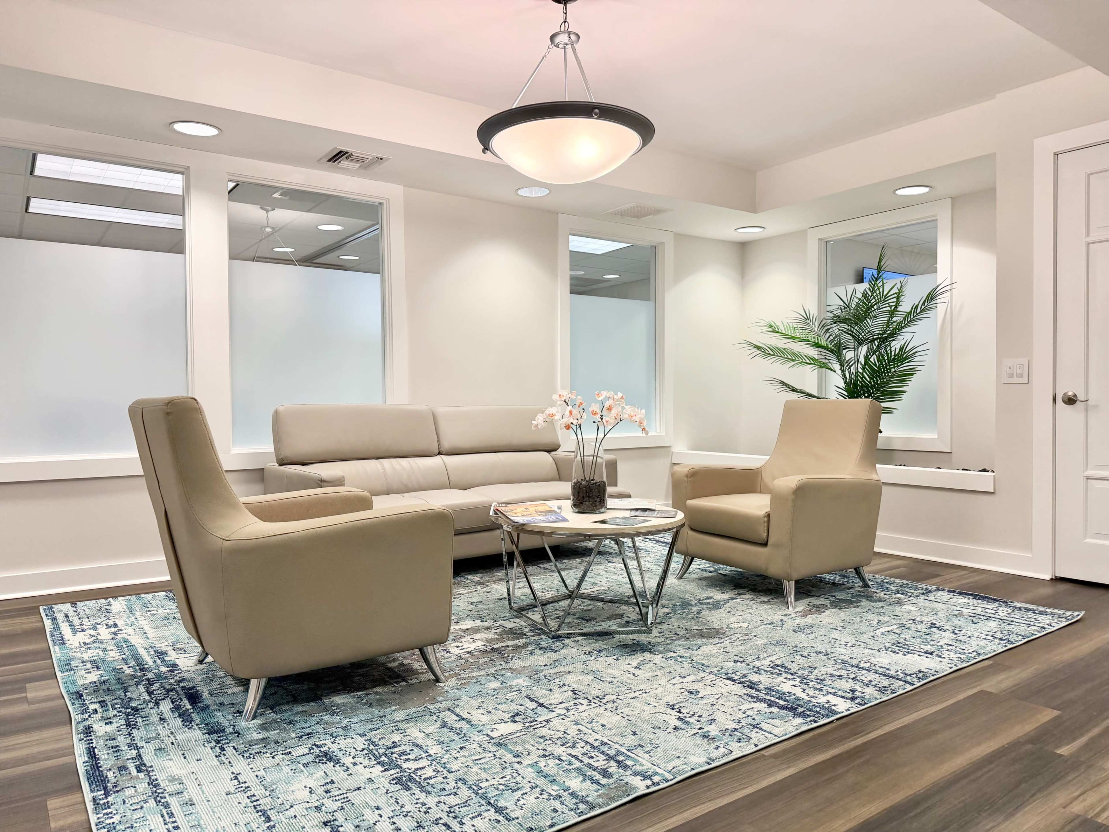 The image shows a modern waiting area featuring a beige sofa, two matching armchairs, a round coffee table with a plant and a floral arrangement, all situated on a blue and white patterned rug.