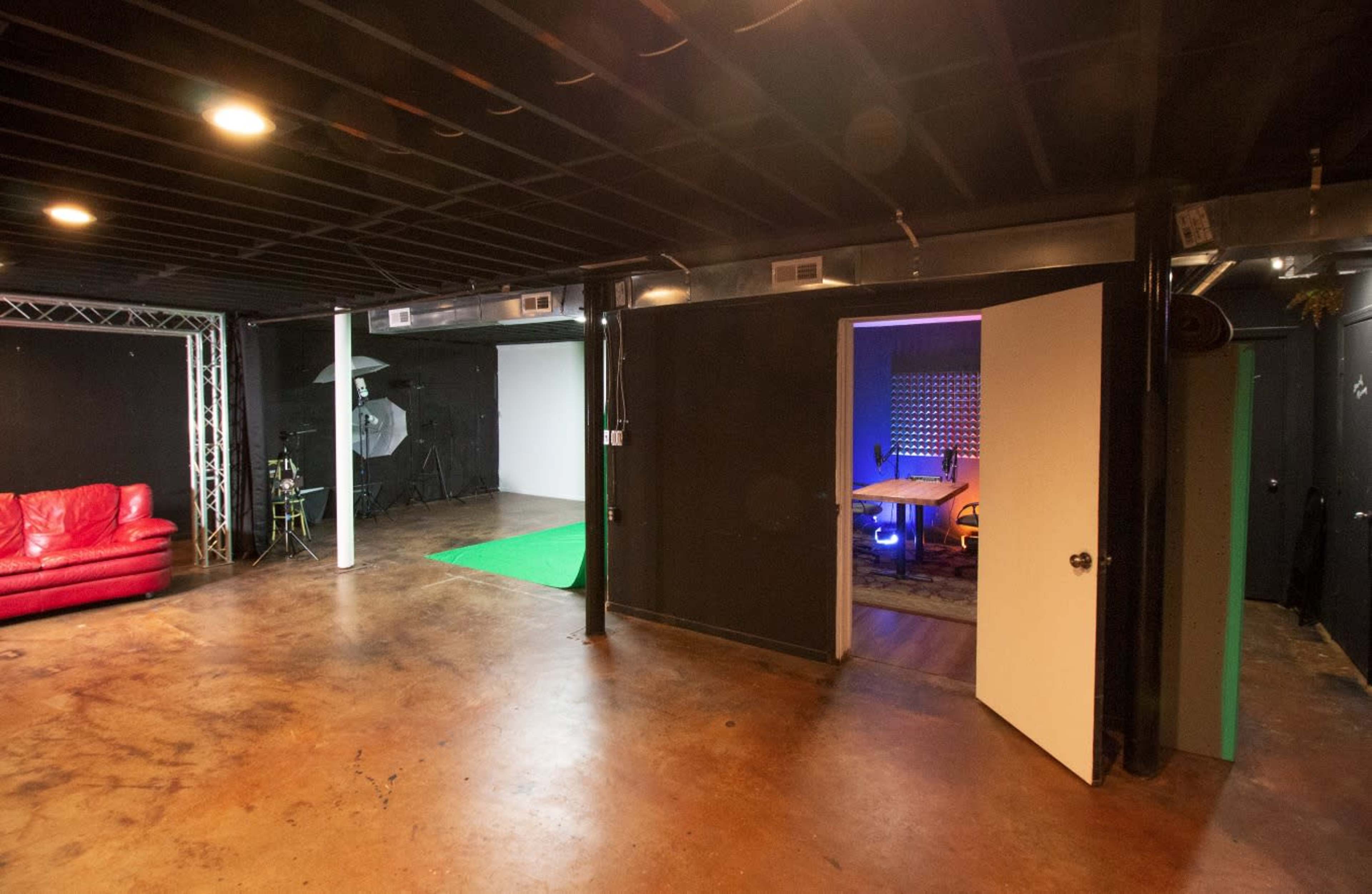 A spacious indoor studio with a green screen area, a red couch, and a doorway leading to a brightly lit room.