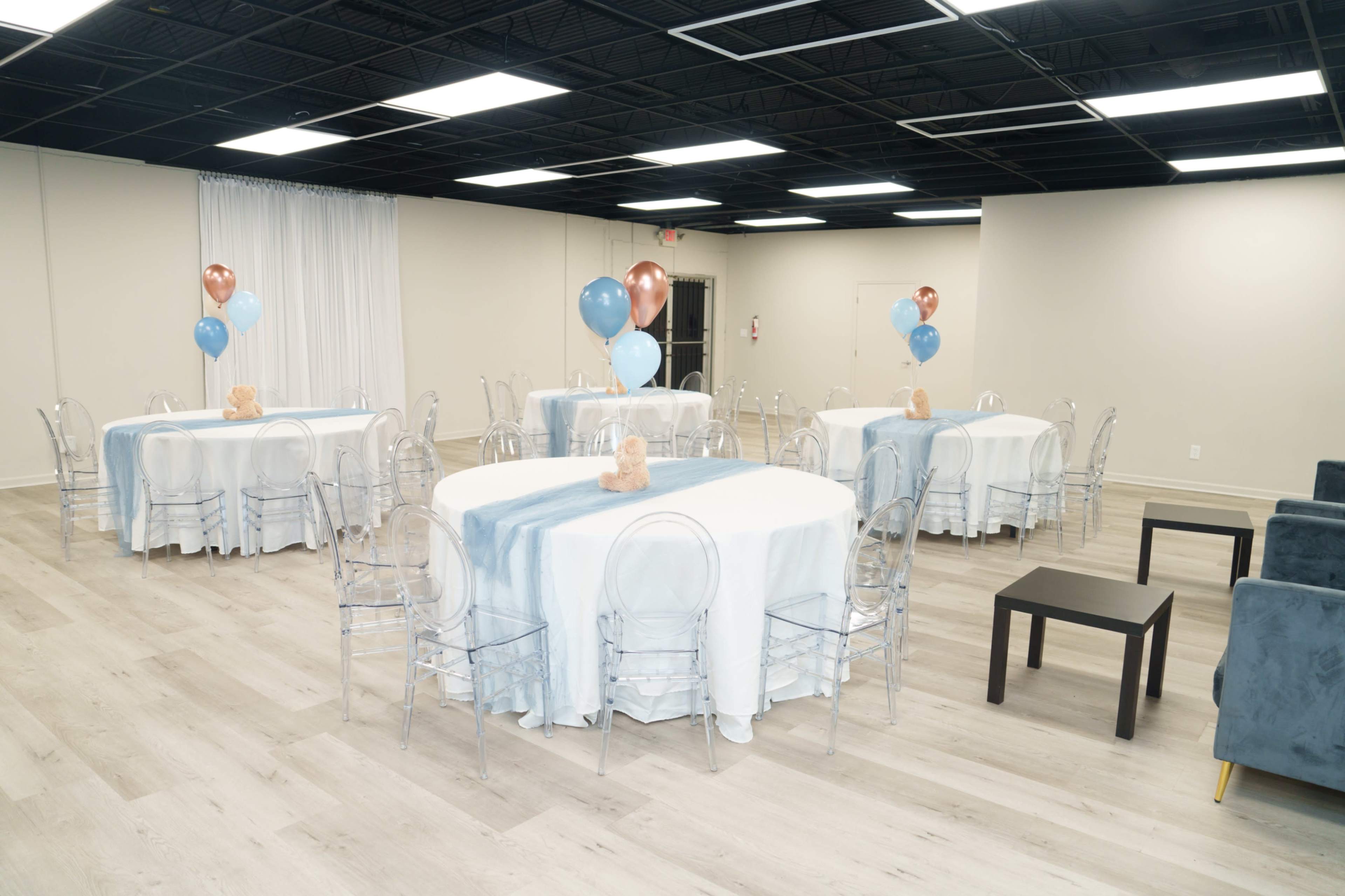 The image shows a spacious event room with six round tables, each covered in light blue and white tablecloths, decorated with balloons in shades of blue and brown.