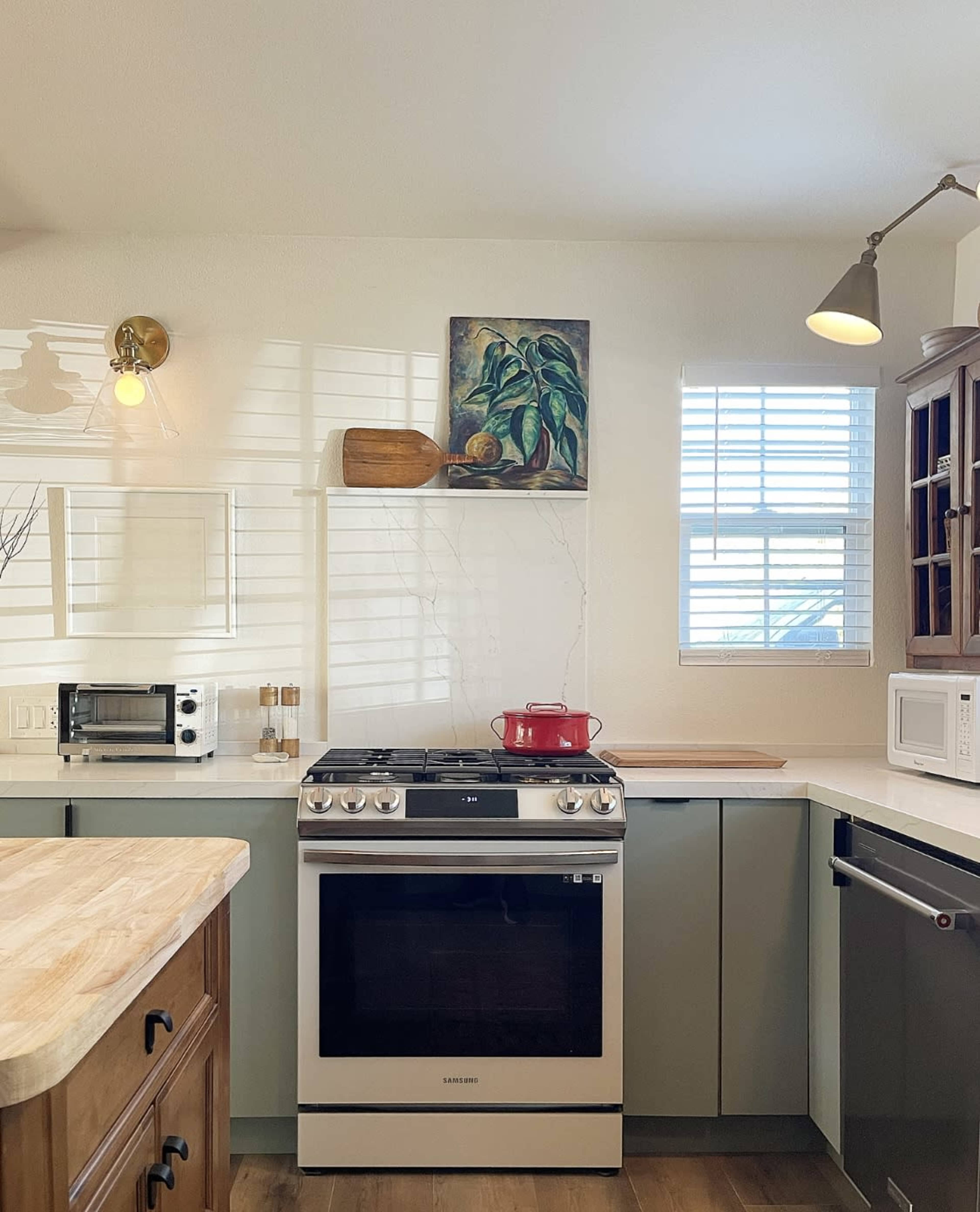 A modern kitchen features a gas stove, a red kettle on the burner, and a small microwave, with a wooden countertop and a painting of a plant on the wall.