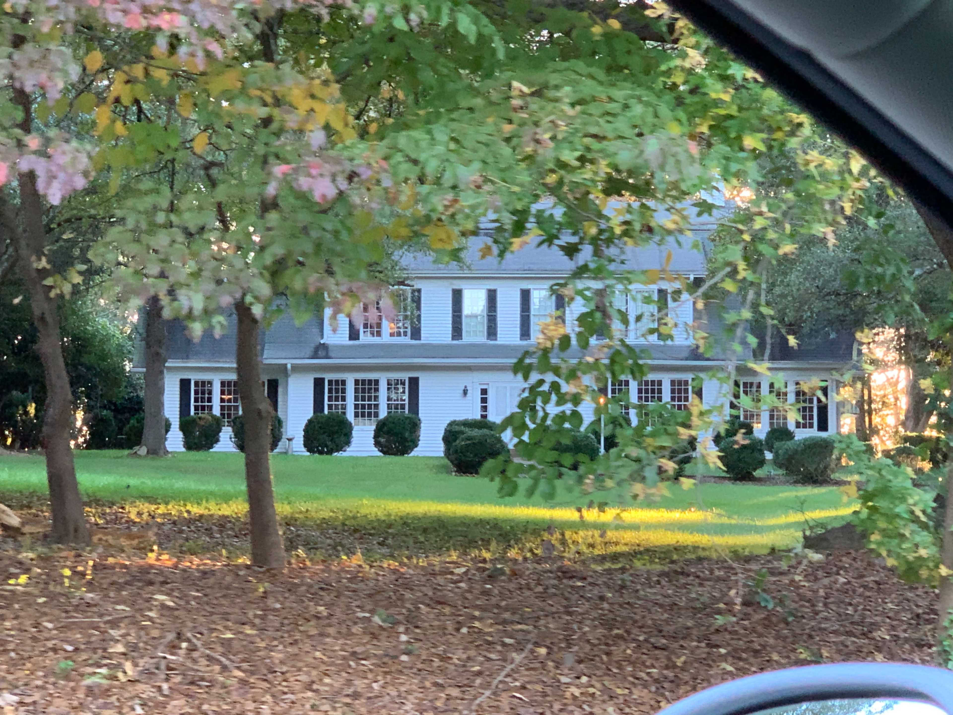 A two-story white house with black shutters is surrounded by trees and a manicured lawn.