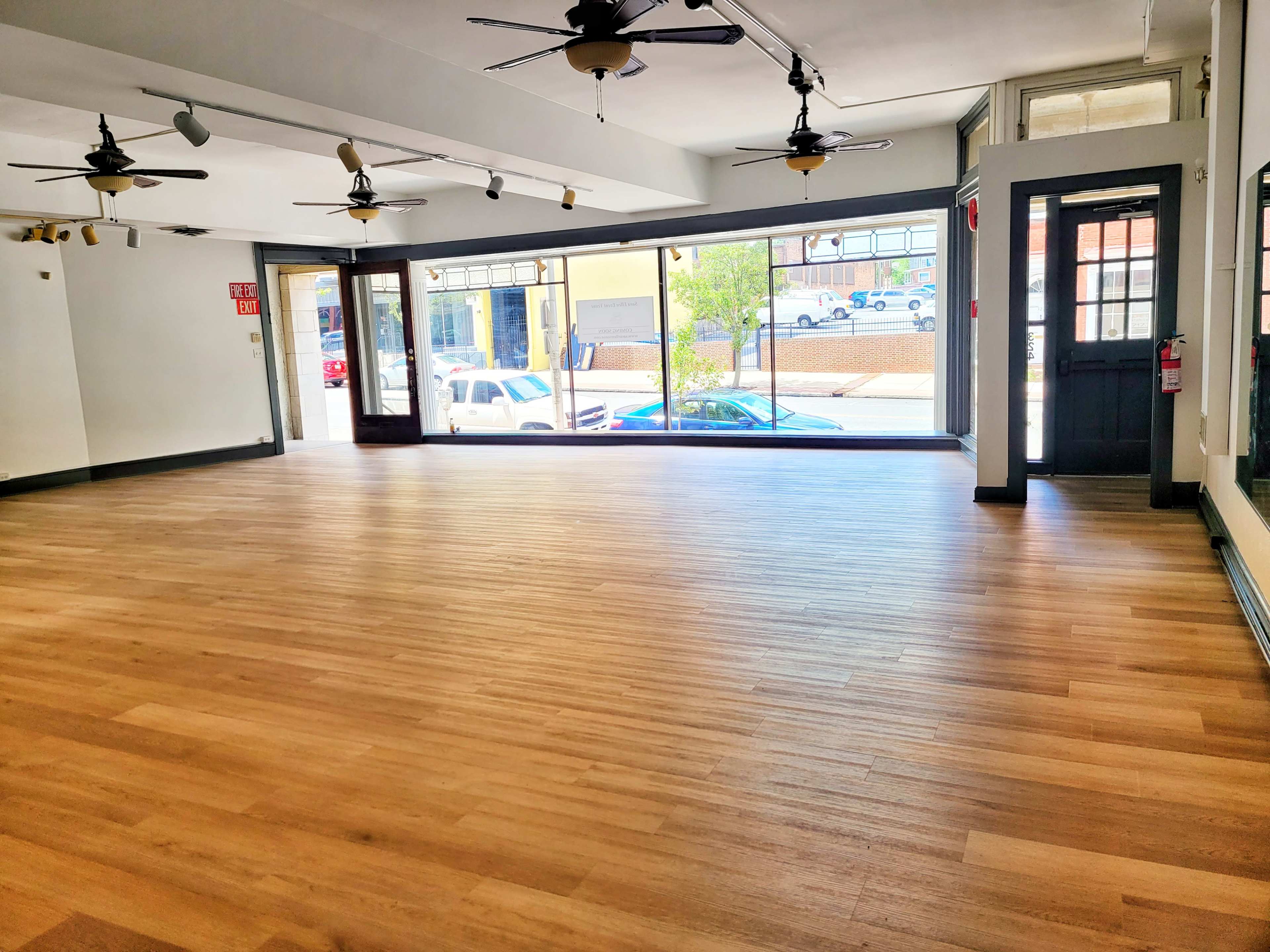 An empty room with wooden floors, large windows, and ceiling fans, allowing natural light to enter.