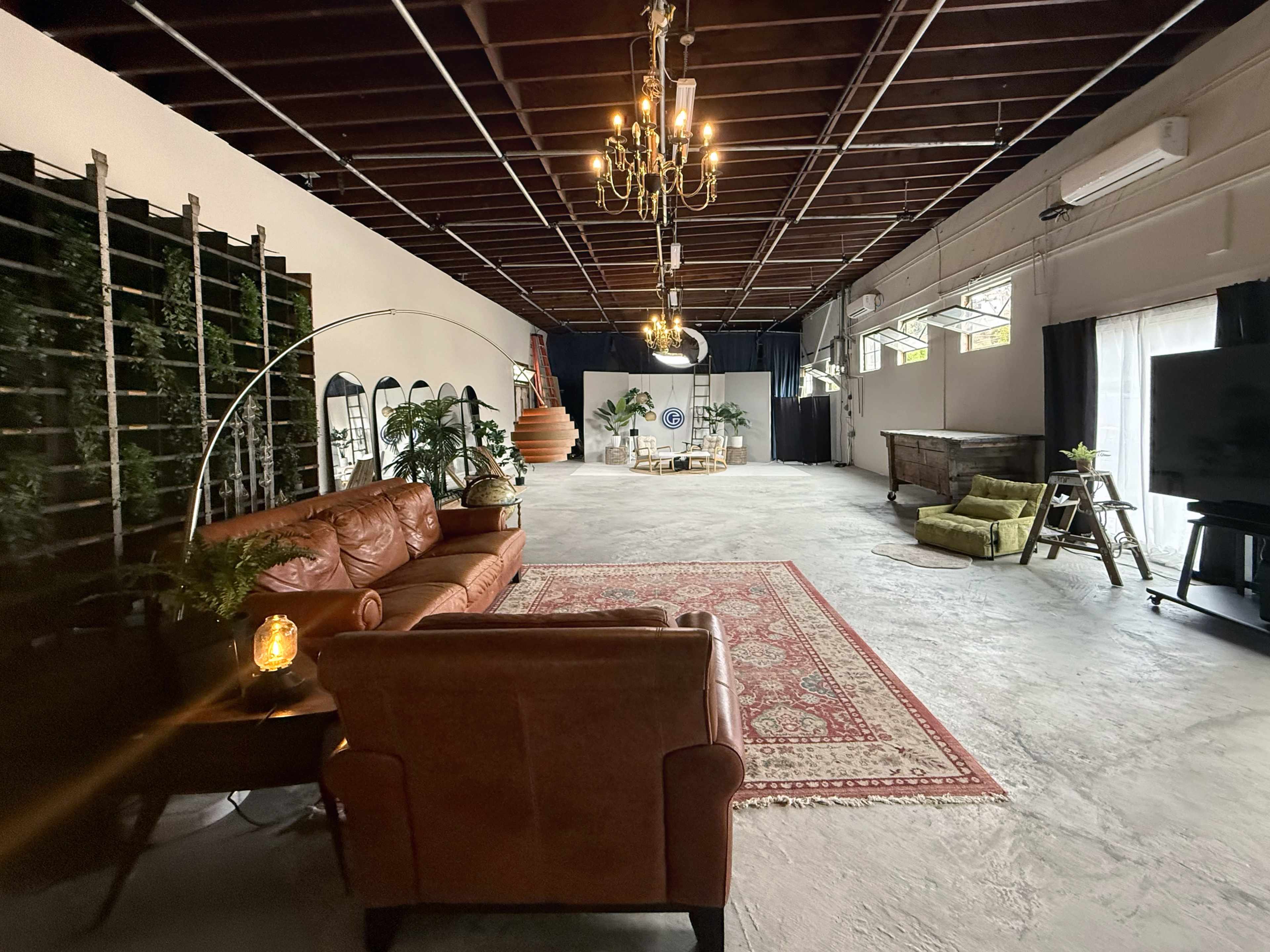 The image shows a spacious indoor area with a leather sofa, greenery, a chandelier, and a large television, featuring a distinctive rug on the concrete floor.