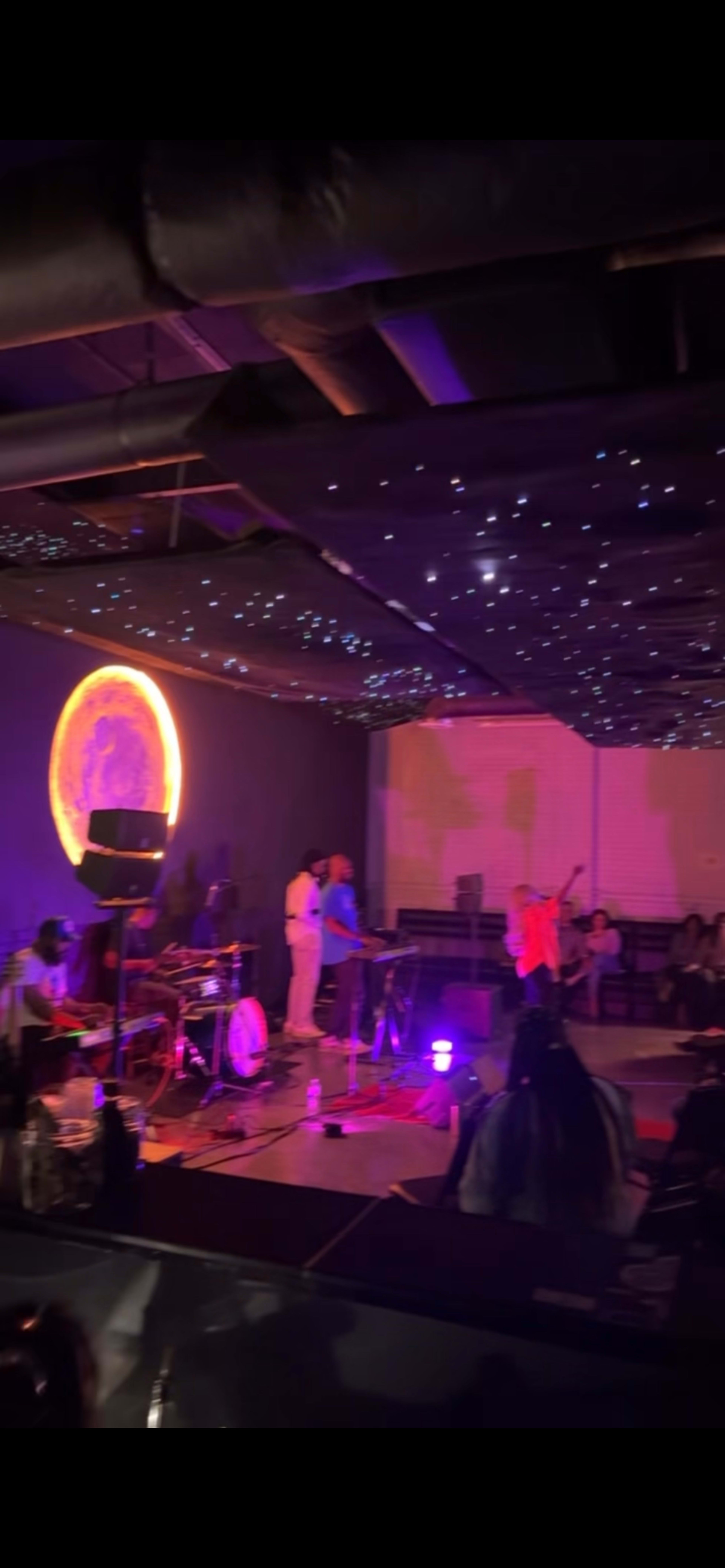 A live music performance takes place under a starry ceiling with a large illuminated moon in the background.