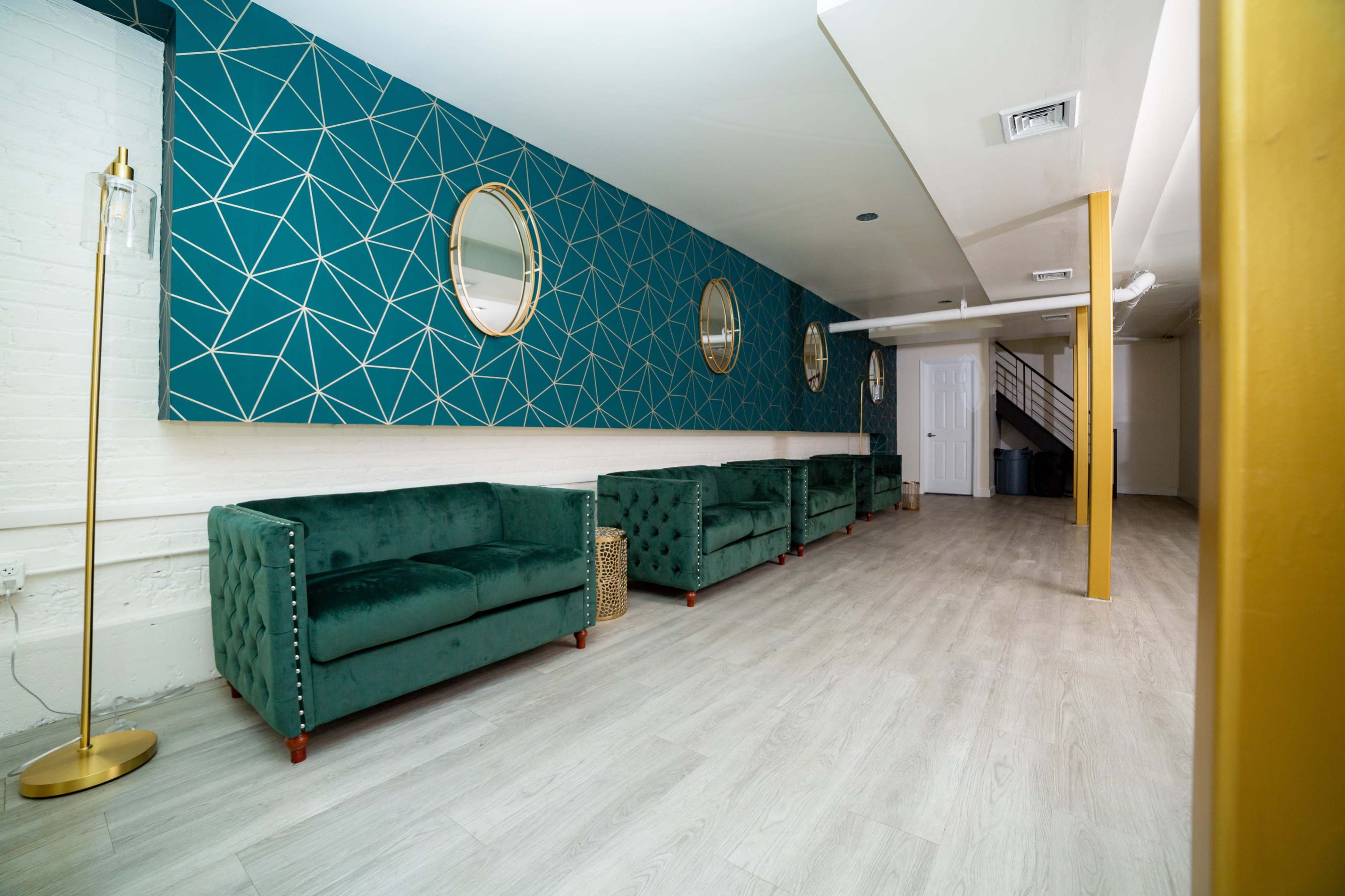 A modern interior space featuring green velvet sofas aligned against a geometric patterned blue wall, with round mirrors and a staircase visible in the background.