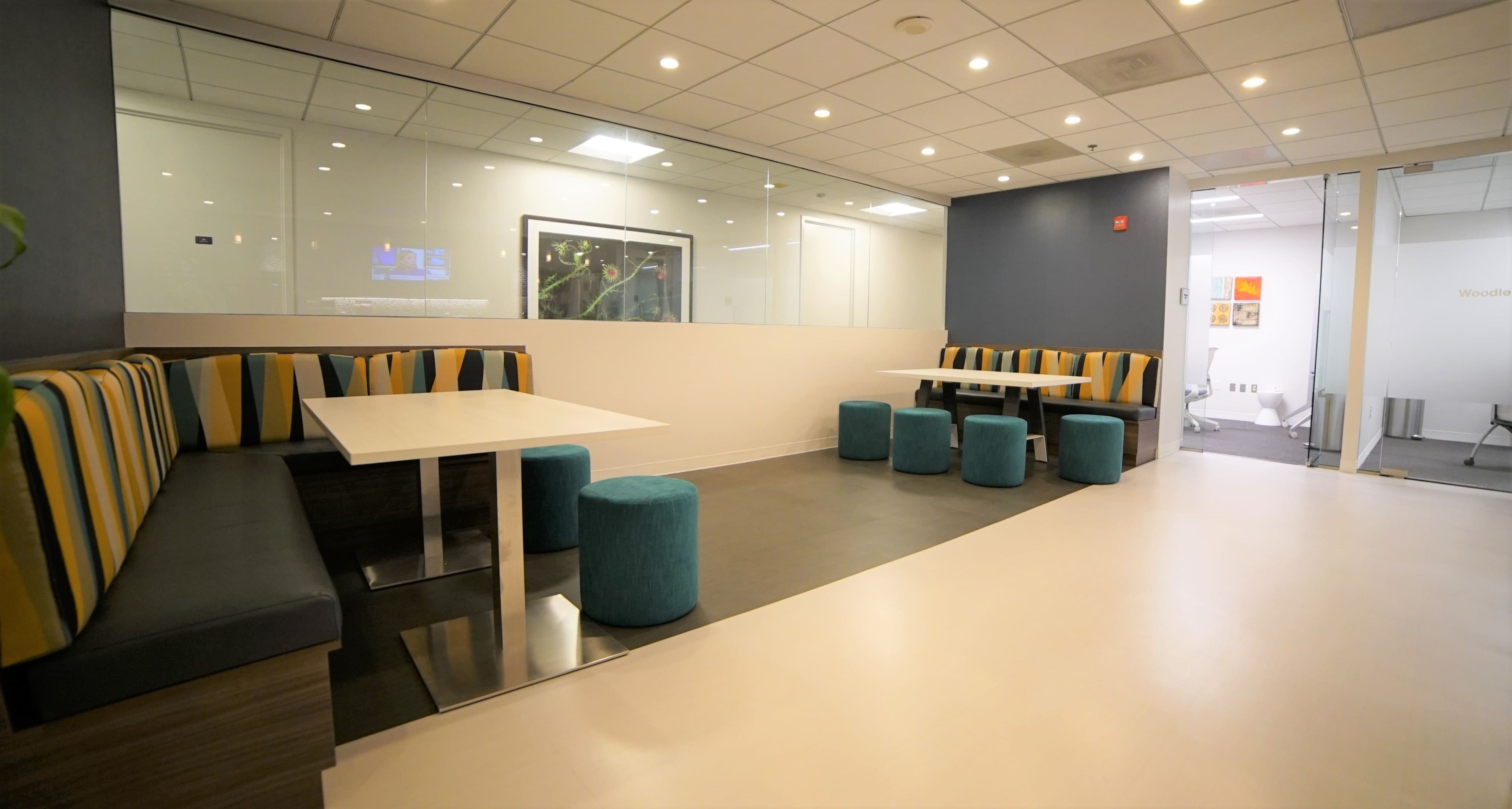 A modern office lounge area with striped seating, blue stools, and partitioned glass walls.