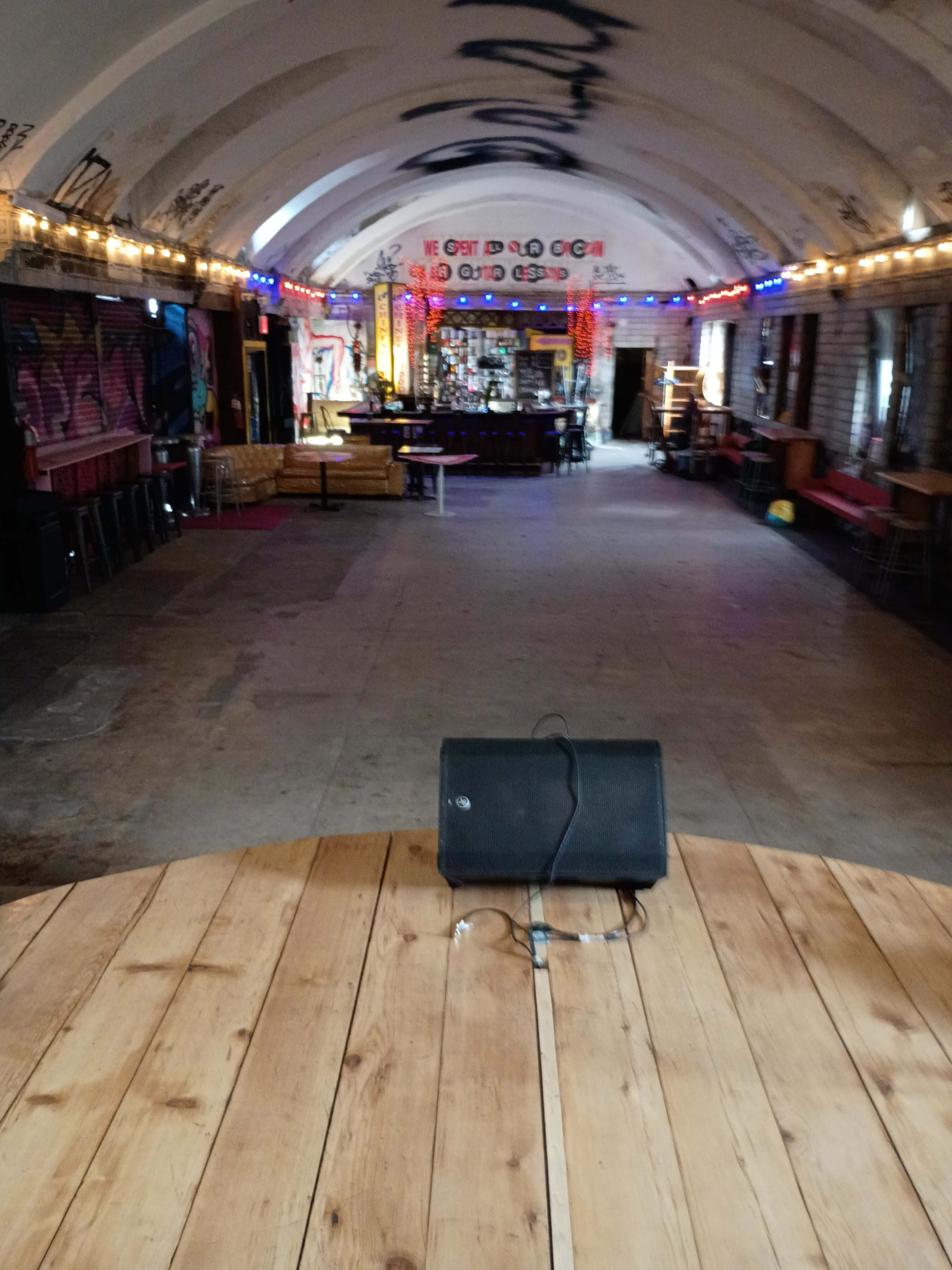 The image shows a spacious, dimly lit venue with colorful wall art, a bar area in the back, and a wooden stage with a speaker facing the audience.