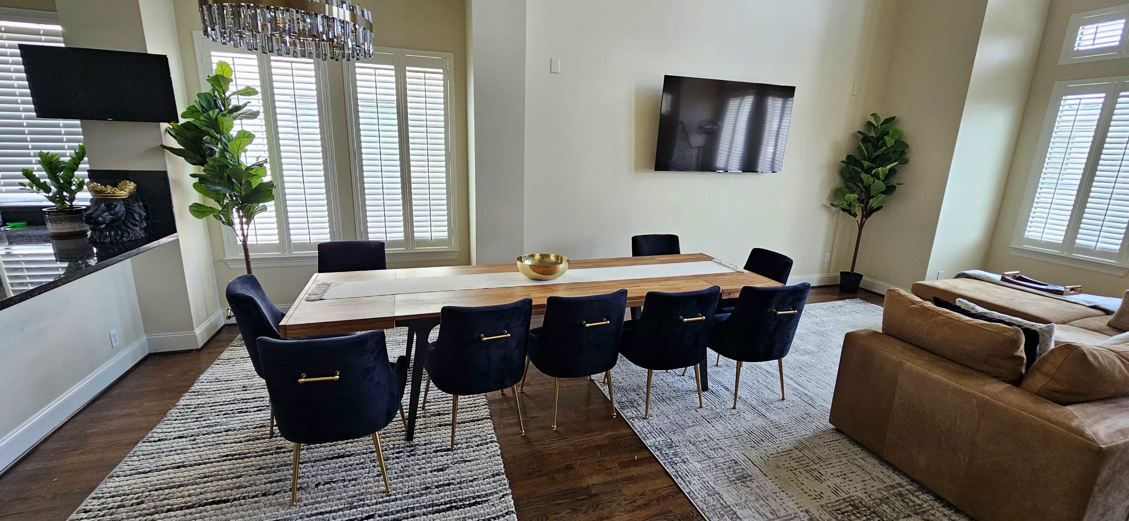 A spacious dining area features a long wooden table surrounded by dark upholstered chairs, with a television mounted on the wall and potted plants in the corners.
