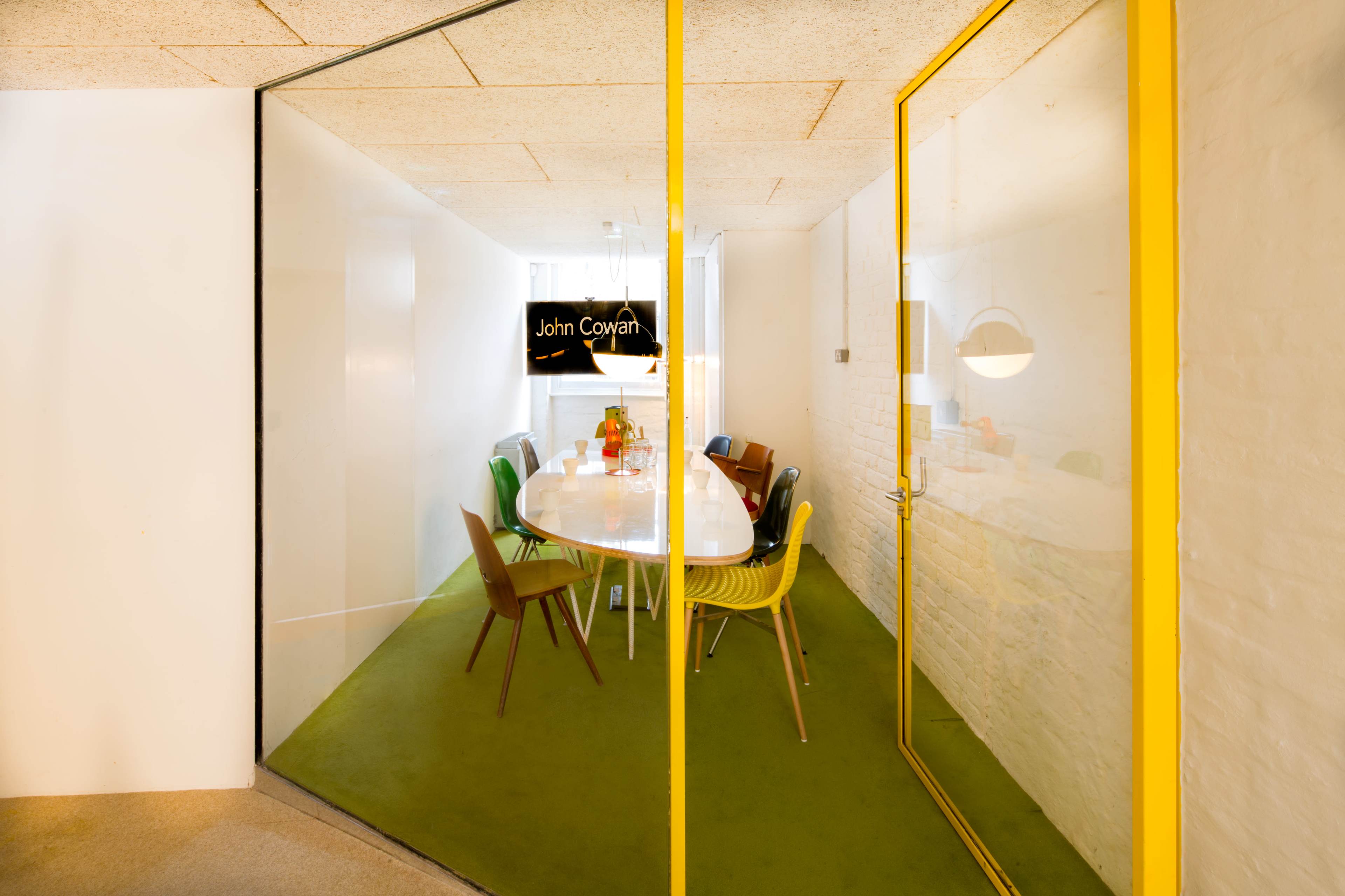 A conference room with a white table surrounded by colorful chairs is visible through glass walls with a yellow door.