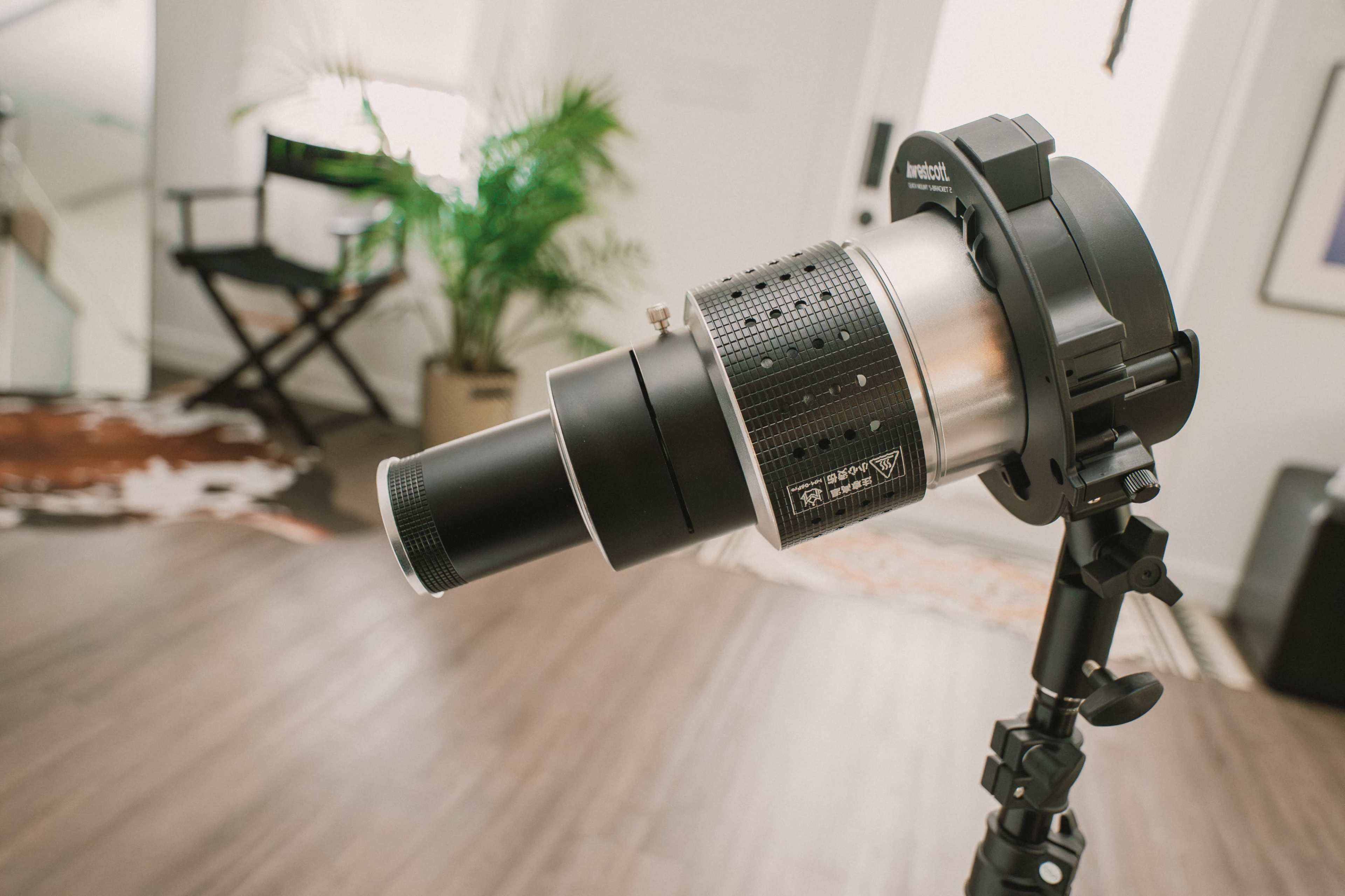 The image shows a close-up of a photography light setup mounted on a stand in a bright room with a large plant in the background.