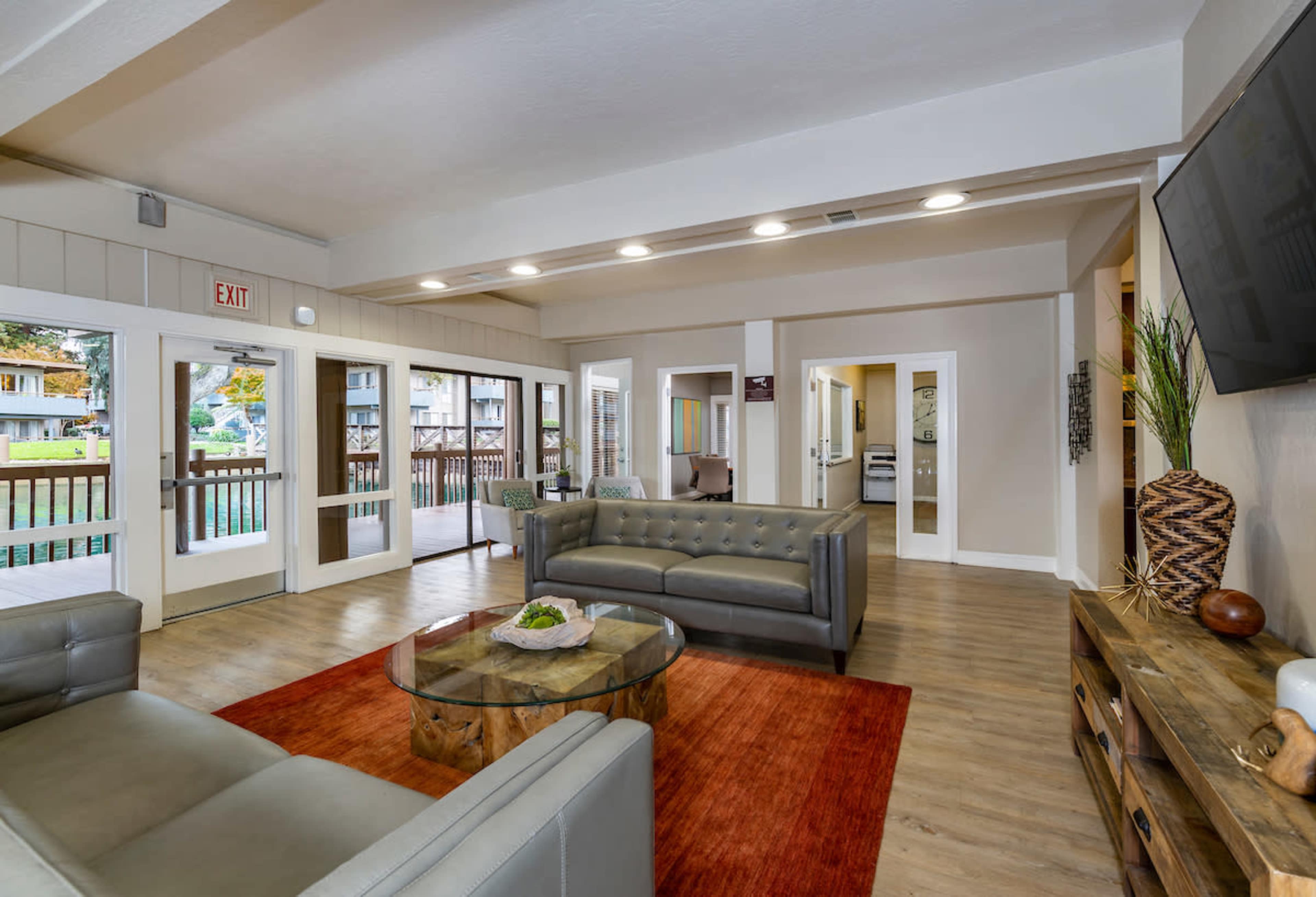 A modern living room featuring gray sofas, a glass coffee table, and large windows allowing natural light, with doors leading to a balcony area.