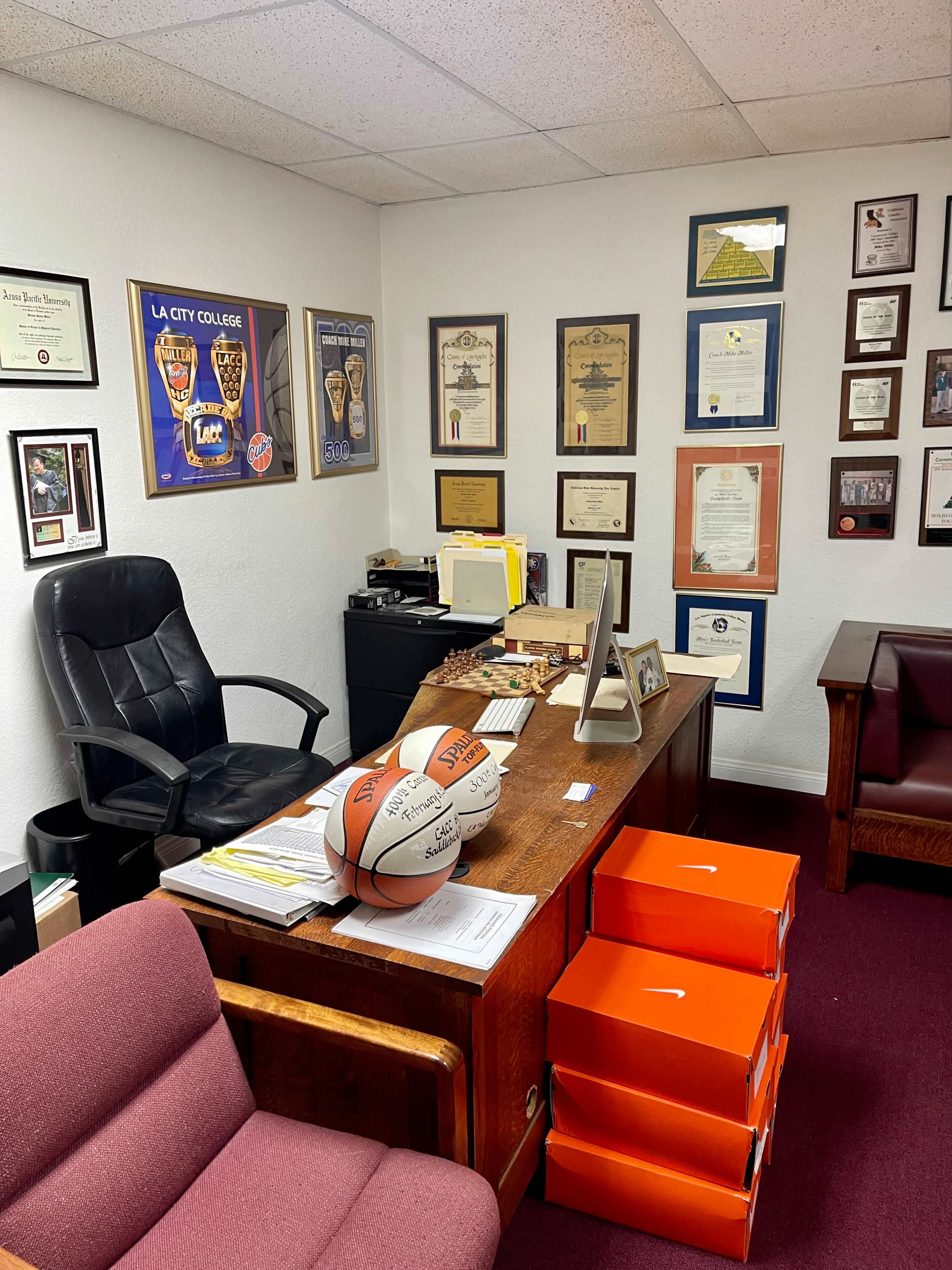 An office with a wooden desk, a black chair, multiple framed certificates and plaques on the walls, and basketballs on the desk alongside orange boxes.