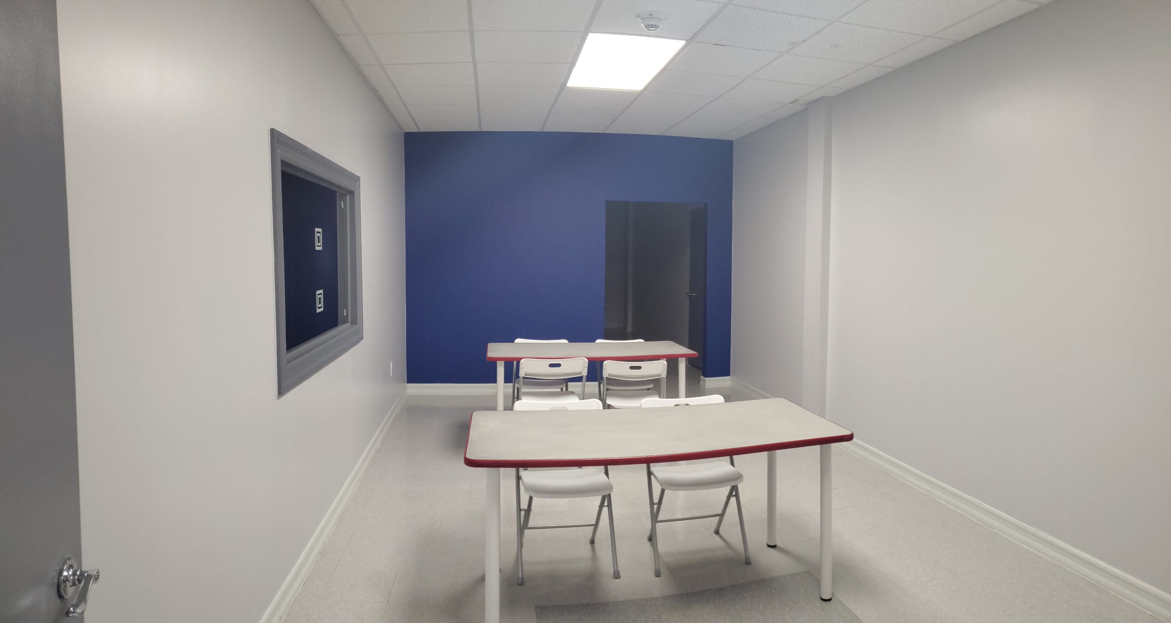 The image shows a small, well-lit classroom with two tables and several white chairs against light gray walls, and a blue accent wall in the background.