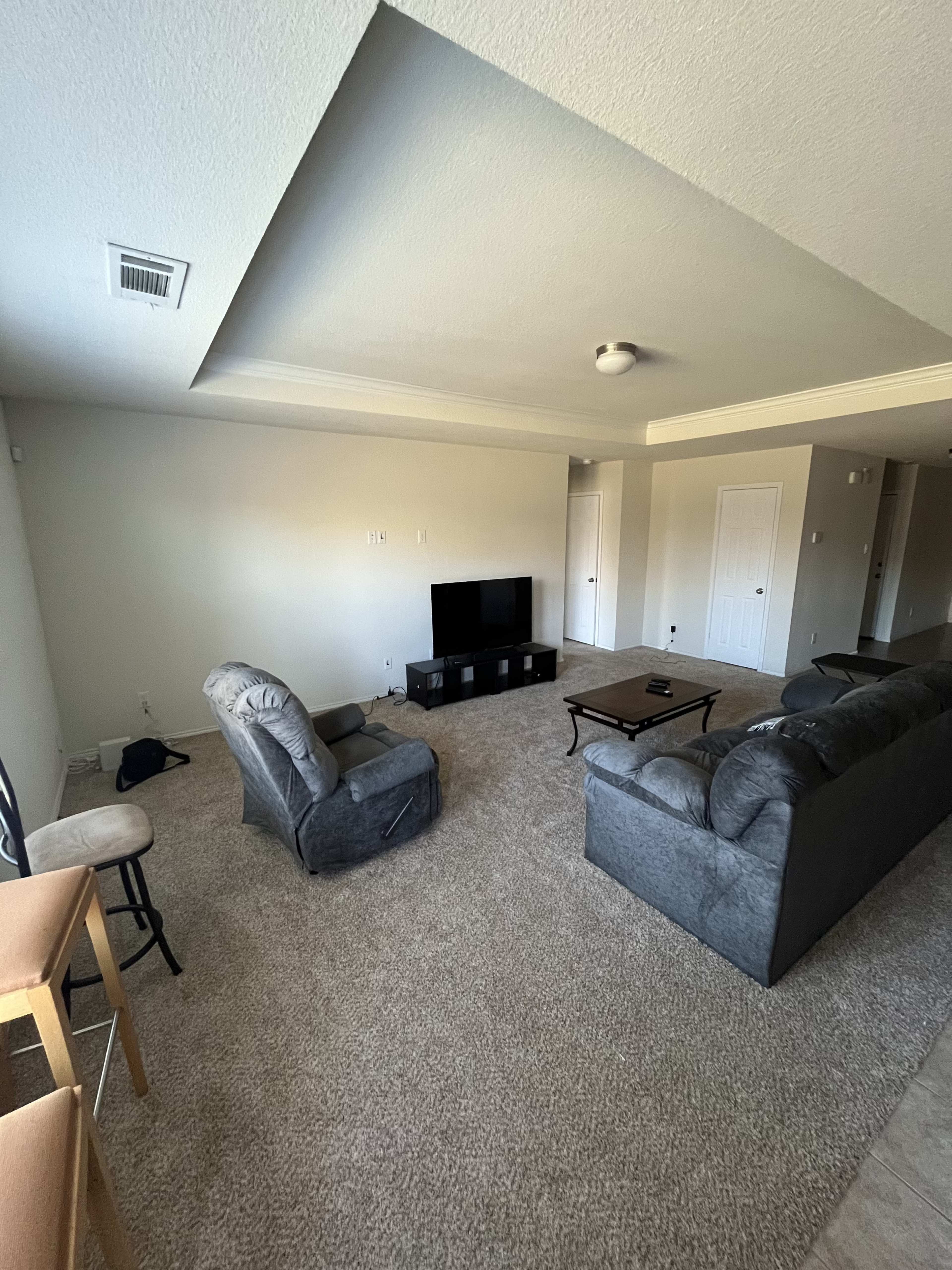 The image shows a spacious living room with a television on a stand, two sofas, a coffee table, and a barstool set against a light-colored wall.