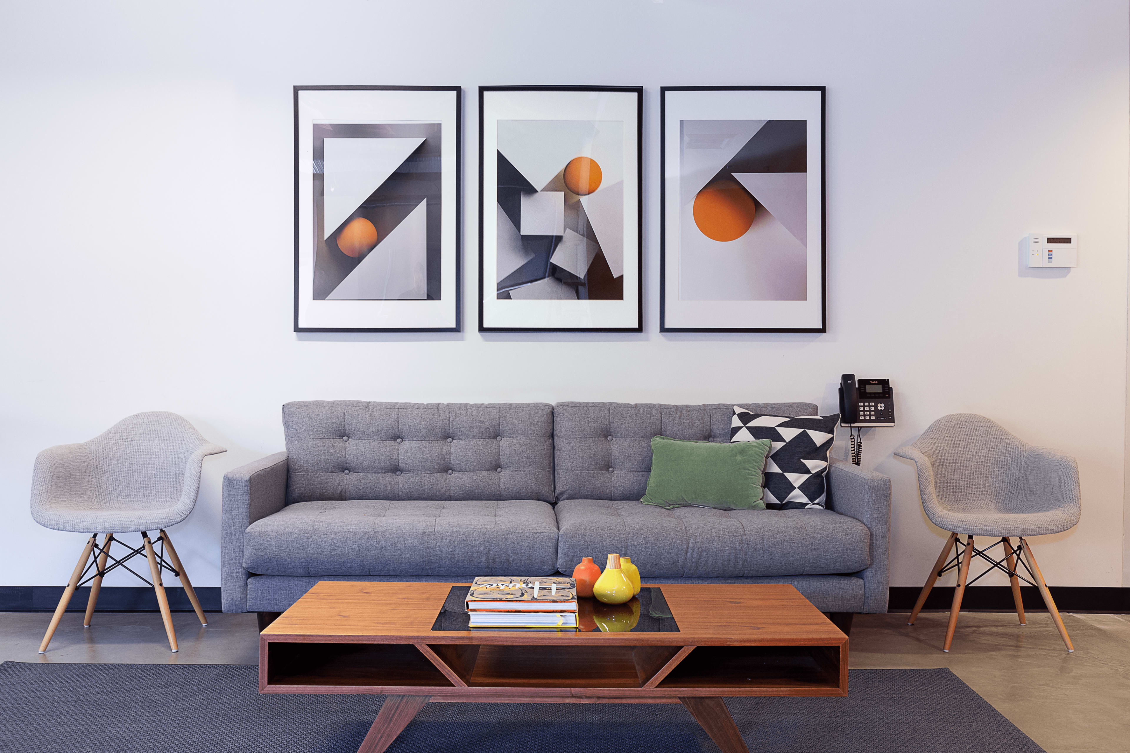 A gray couch with green and patterned cushions sits in front of a wooden coffee table, flanked by two gray chairs, with three framed abstract artworks above it.