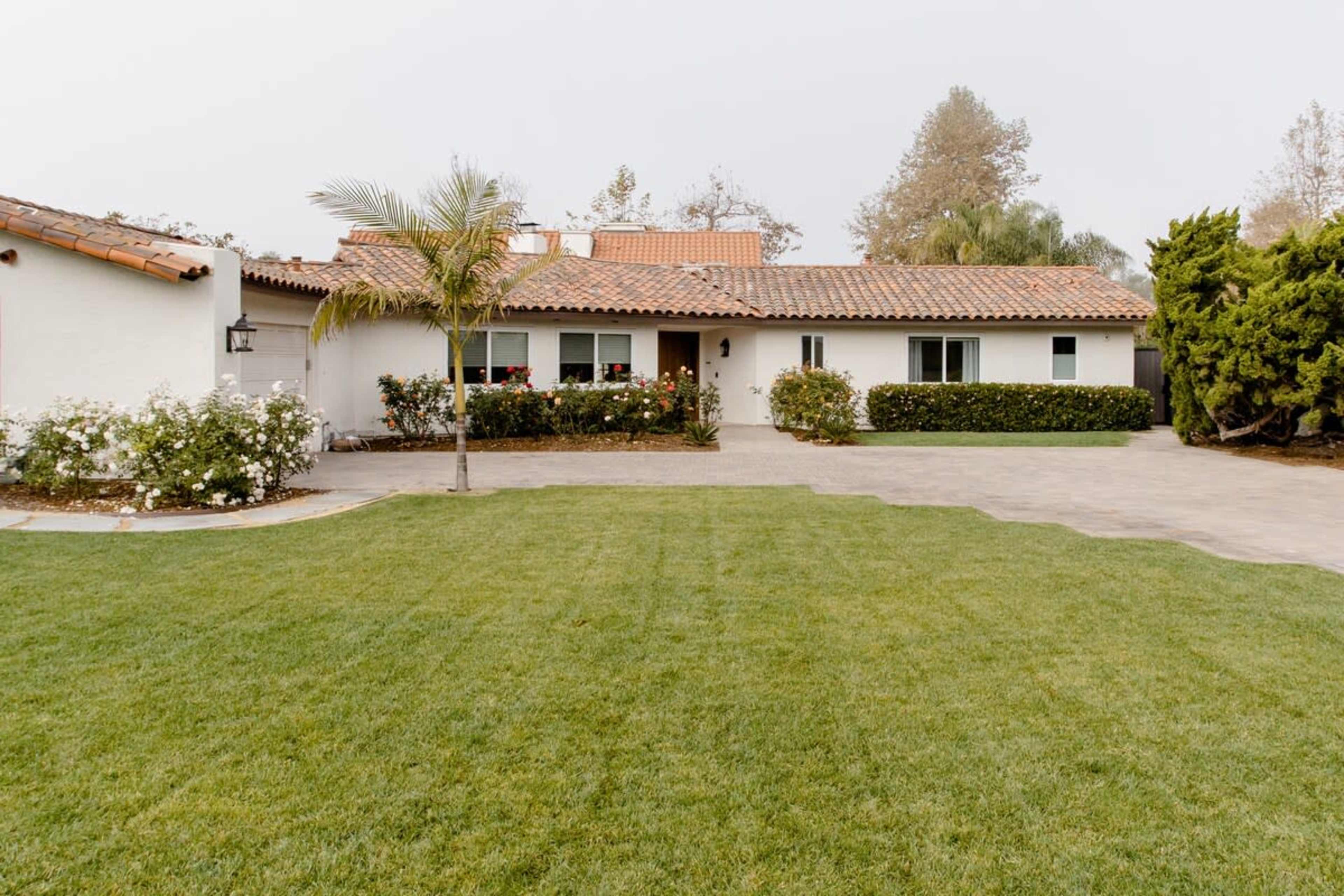 A one-story house with a tiled roof is set behind a manicured lawn and surrounded by shrubbery and flowers.