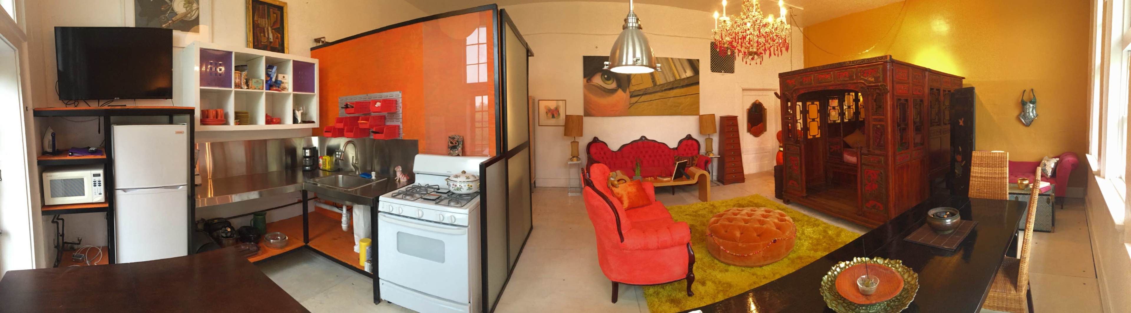 A panoramic view of a stylish living space featuring a kitchen with modern appliances, a vibrant orange accent wall, a cozy seating area with red furniture, and a decorative wooden cabinet.