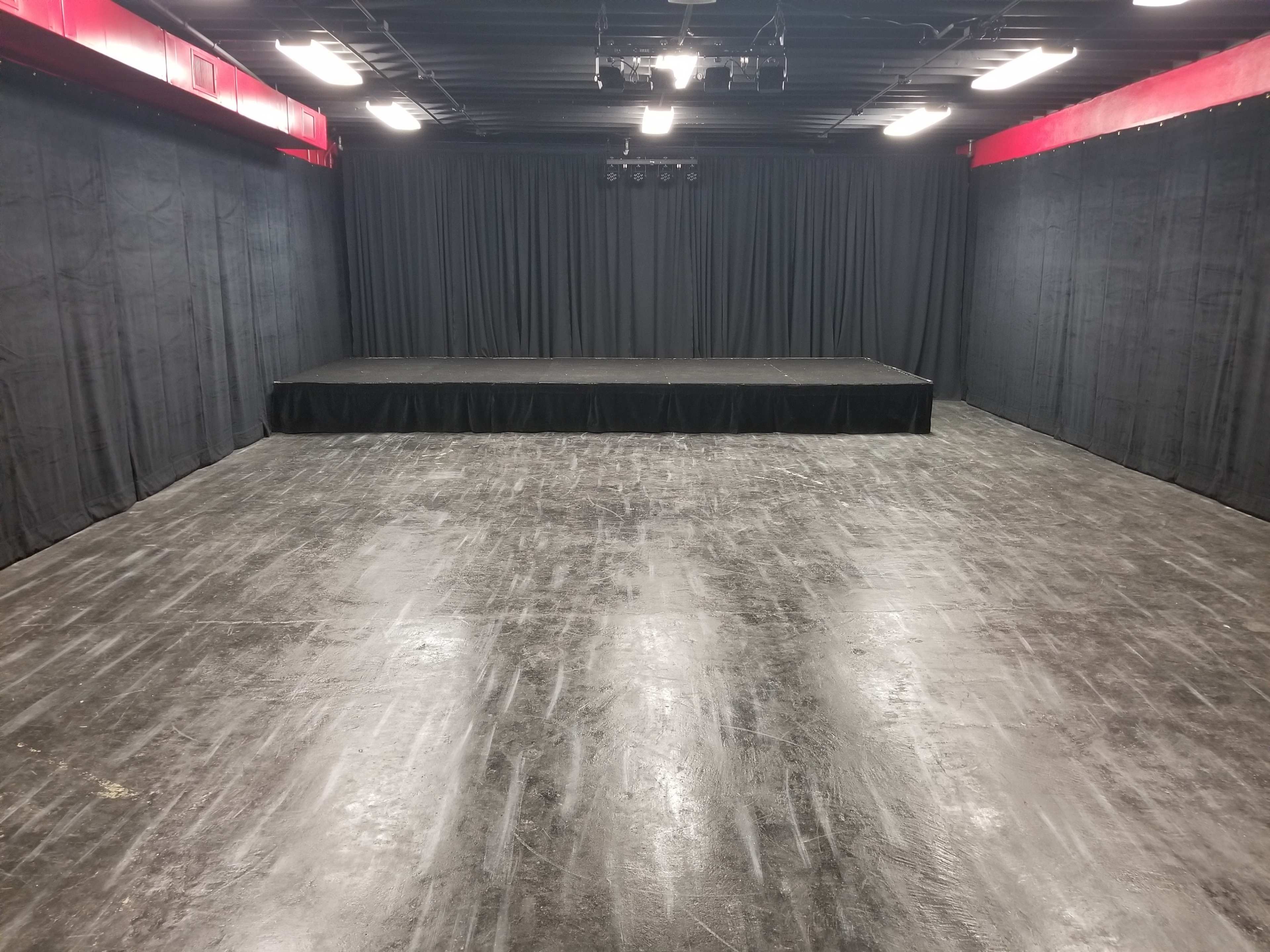 The image shows a dark, empty stage area with black curtains, a raised platform in the center, and a polished concrete floor.