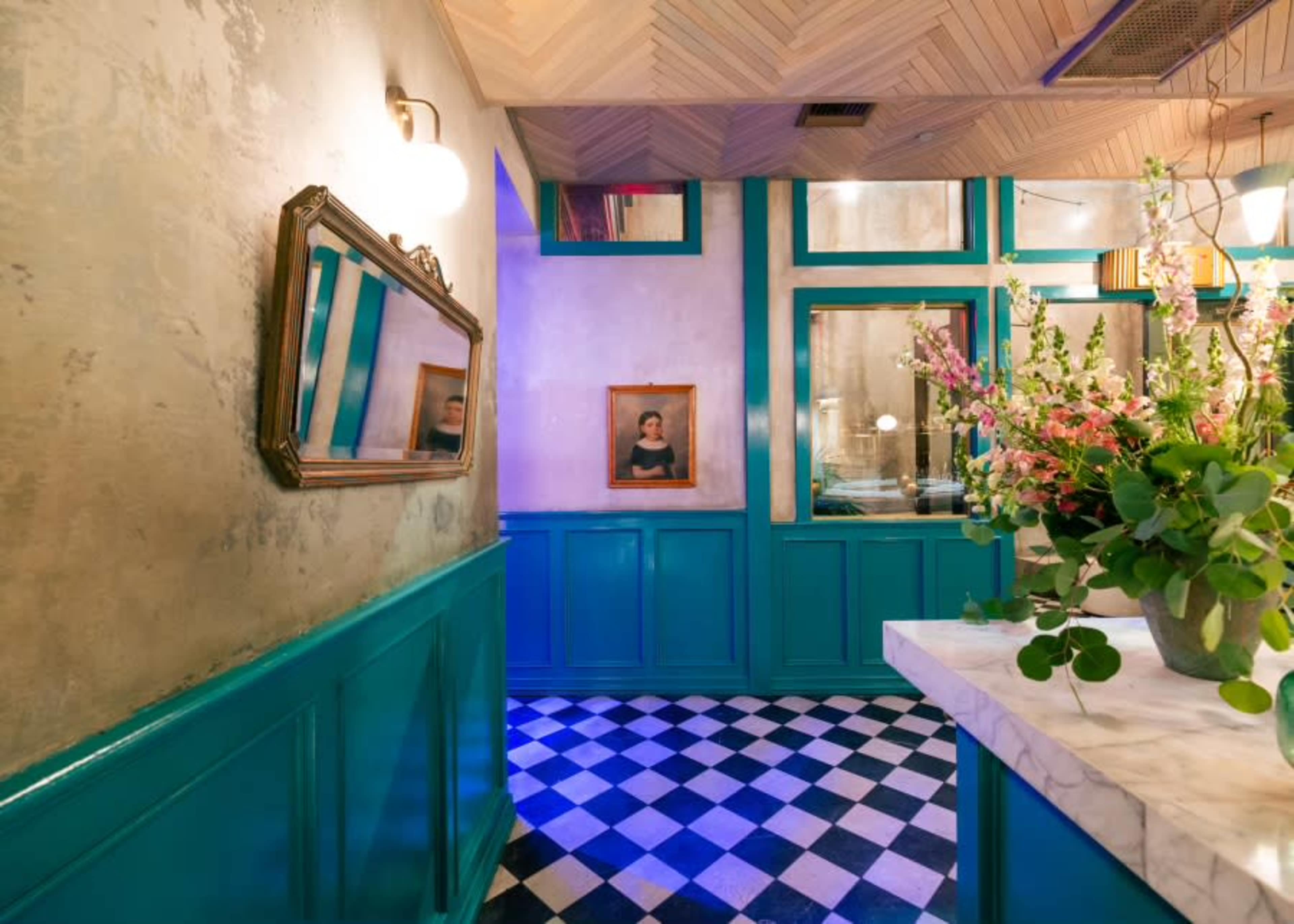 The image shows a corridor with a checkered floor, teal walls, framed portraits, and a marble counter adorned with flowers.