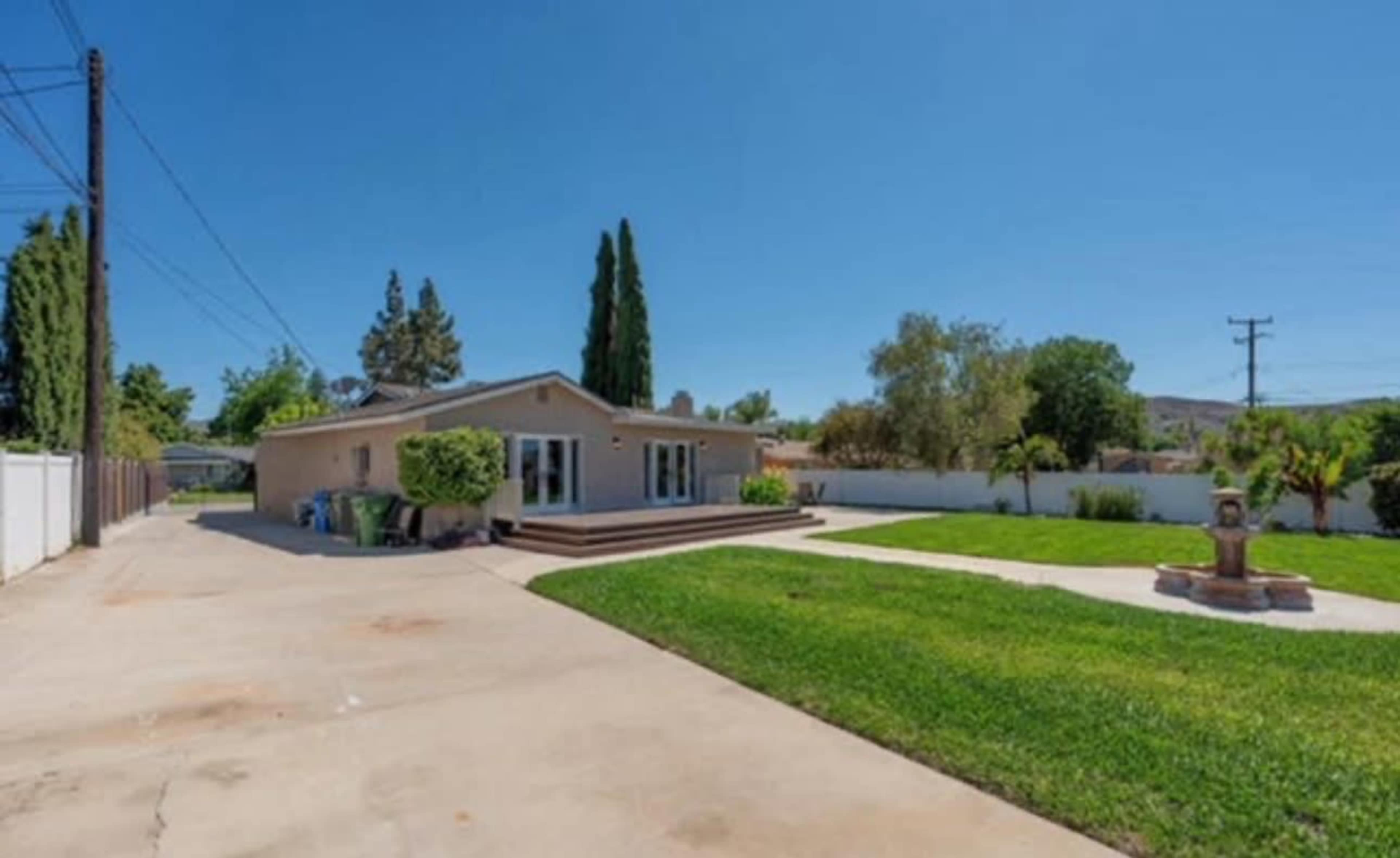 The image shows a single-story house surrounded by grassy areas and trees, with a concrete driveway leading up to it.