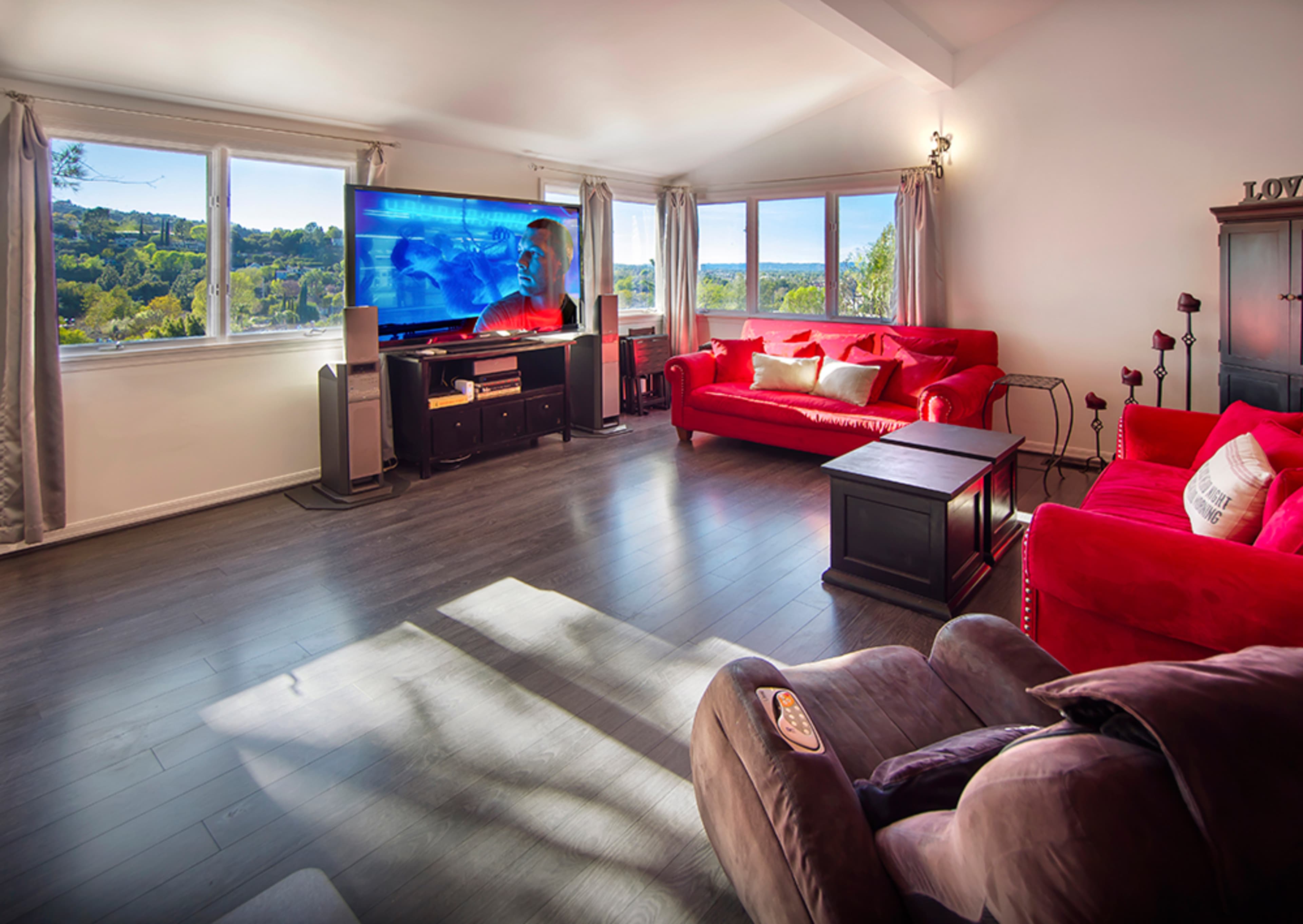 A spacious living room features large windows, red sofas, a dark coffee table, and a television mounted on a cabinet.