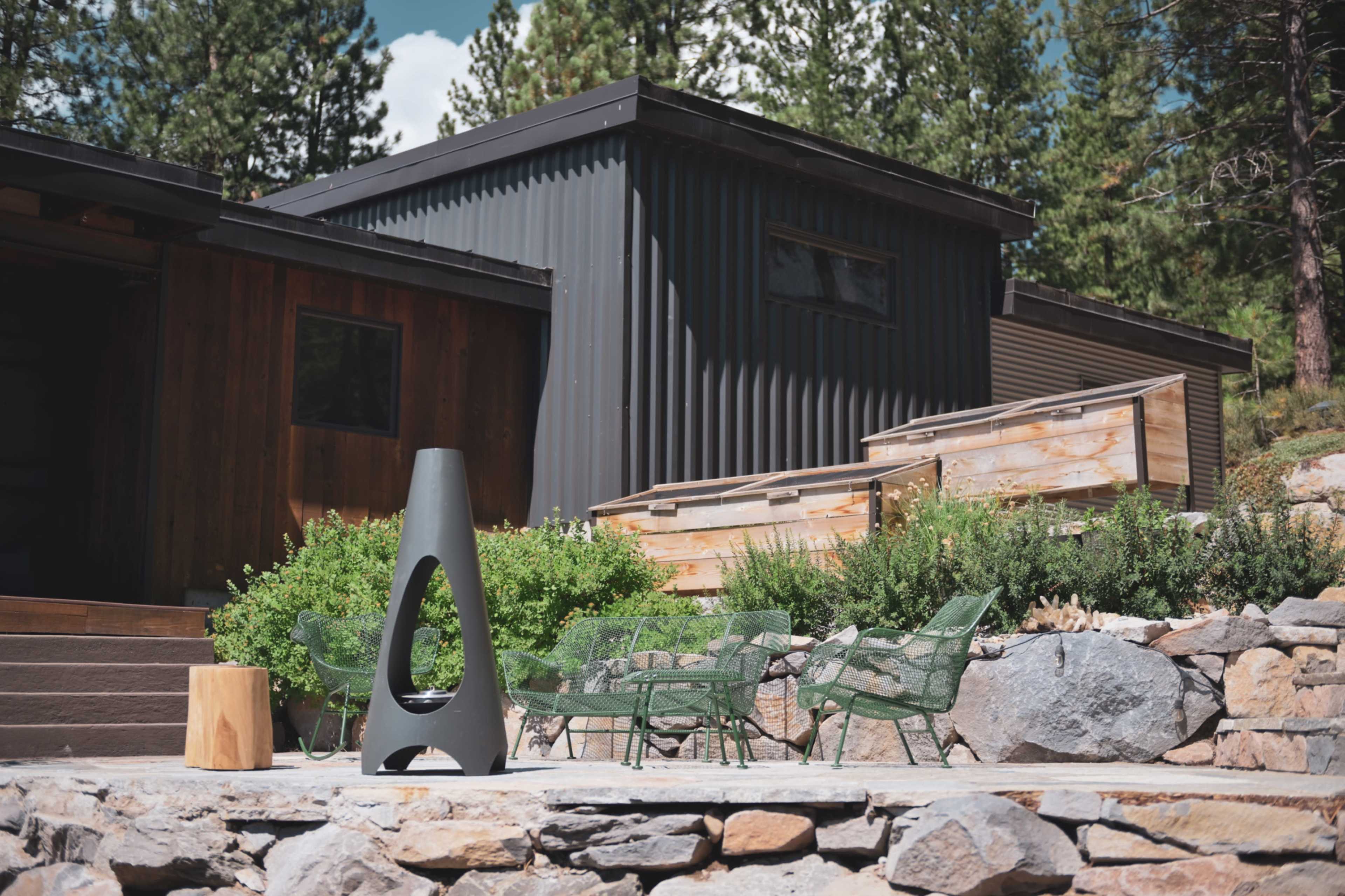A modern outdoor area with a charcoal-colored fire pit, green chairs, and wooden planters, set against a backdrop of two contemporary buildings and trees.