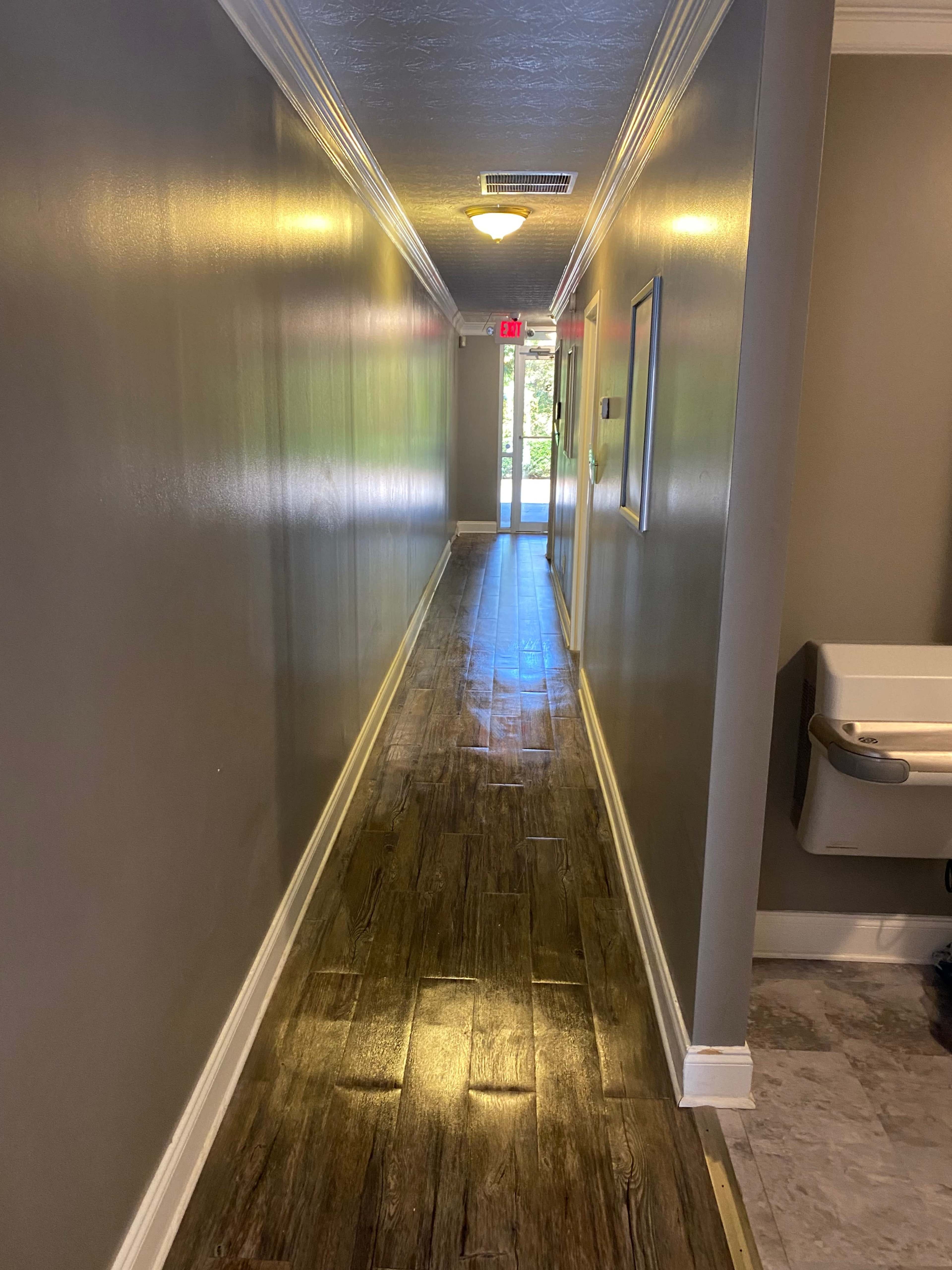 A long, narrow hallway with a wooden floor and walls painted in a neutral color, leading to a glass door that opens to the outside.