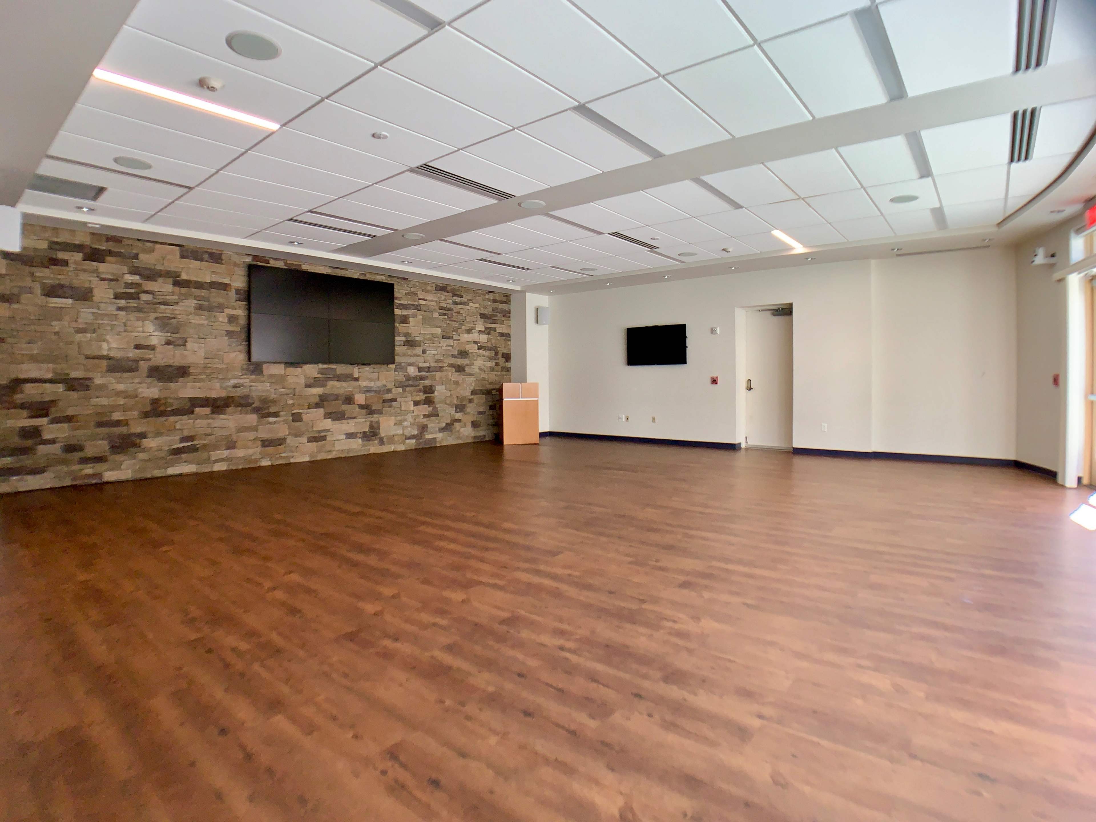 A large, empty room with wooden flooring, a stone accent wall, and two mounted screens on the walls.